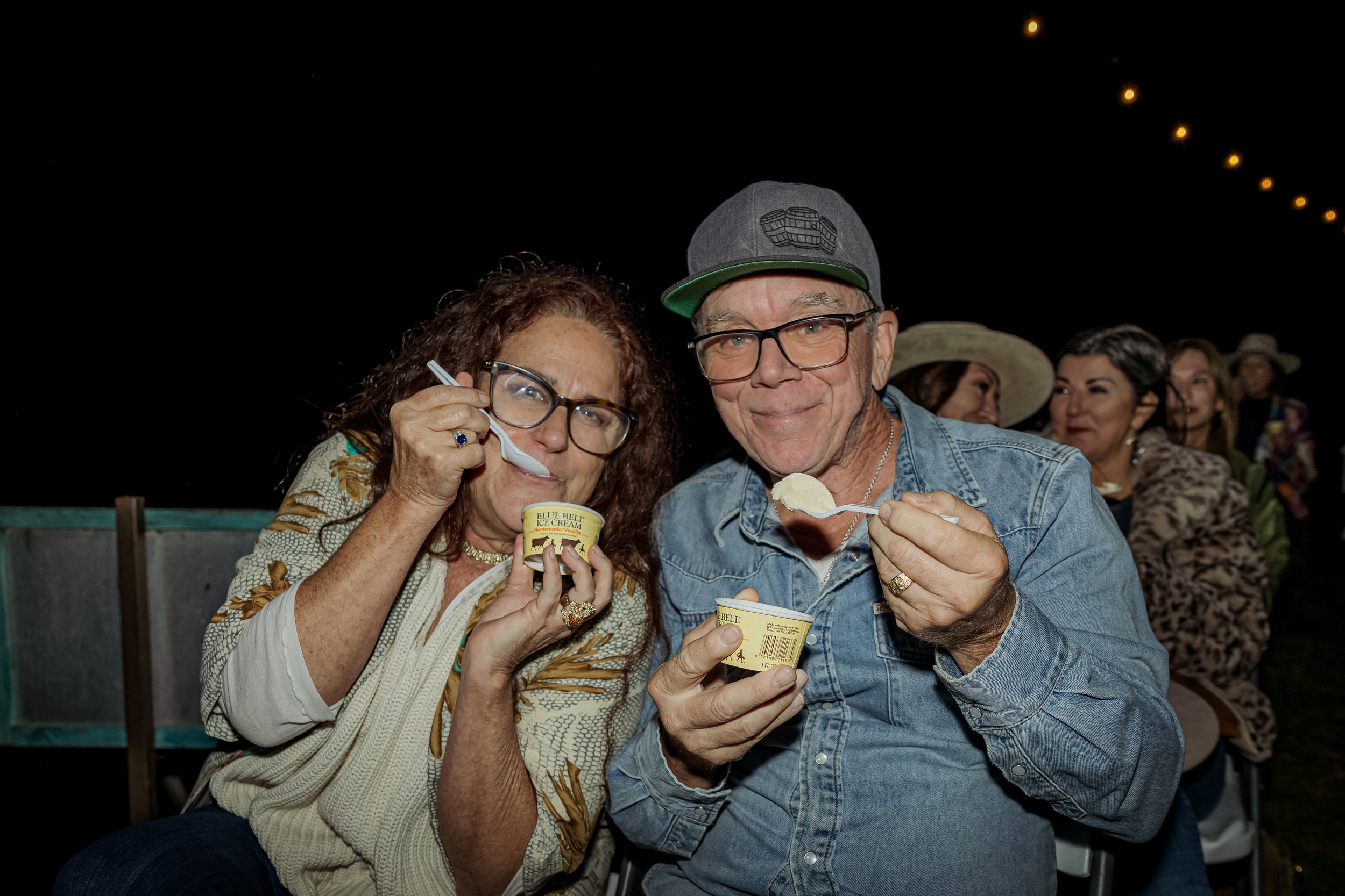 Two older adults enjoying ice cream at night, with other people sitting in the background, under string lights.