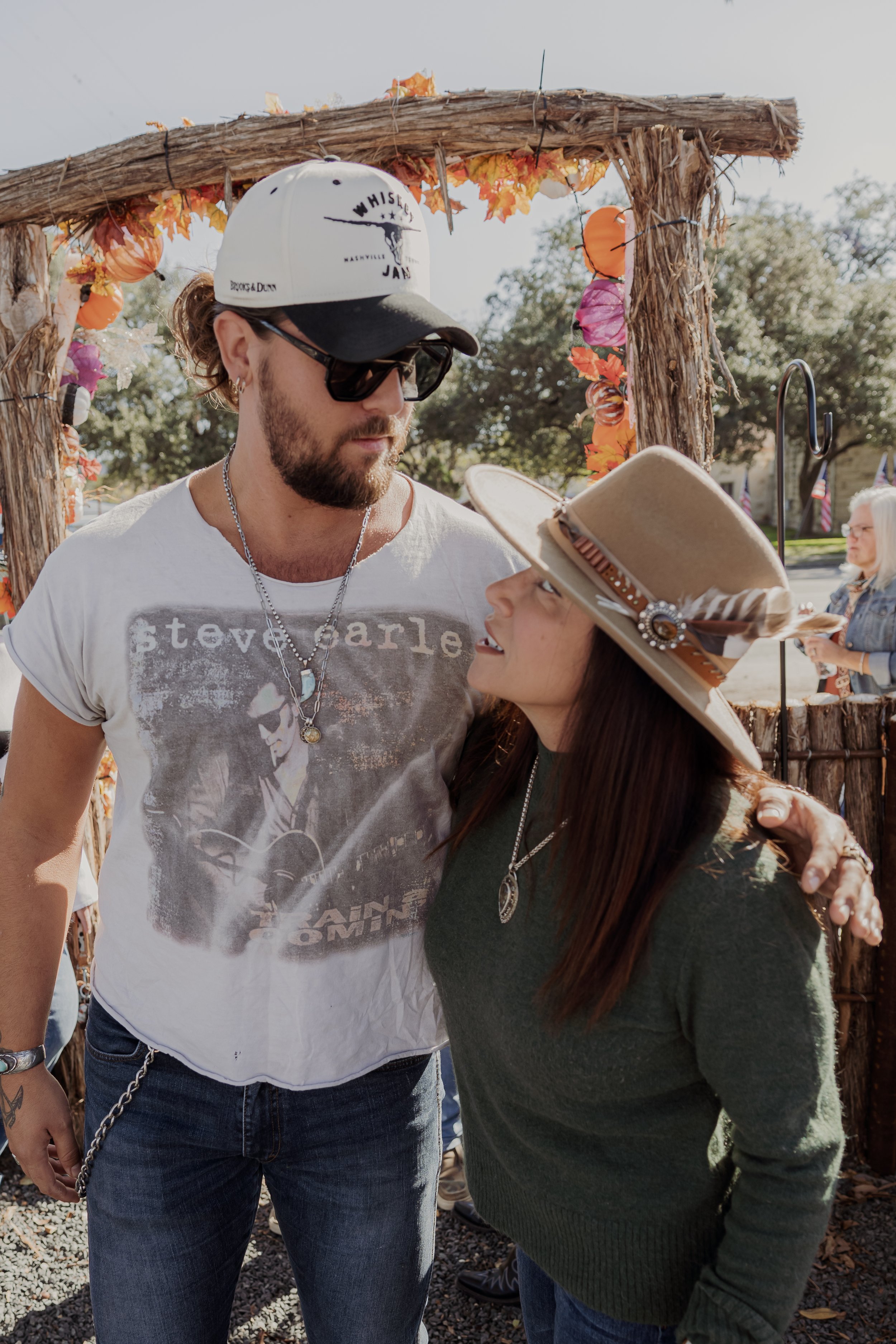 A man and woman are standing close together outdoors, looking into each other's eyes. The man wears sunglasses, a white cap, a Steve Earl t-shirt, and a chain necklace. The woman wears a wide-brimmed tan hat with a feather and a green sweater. They a