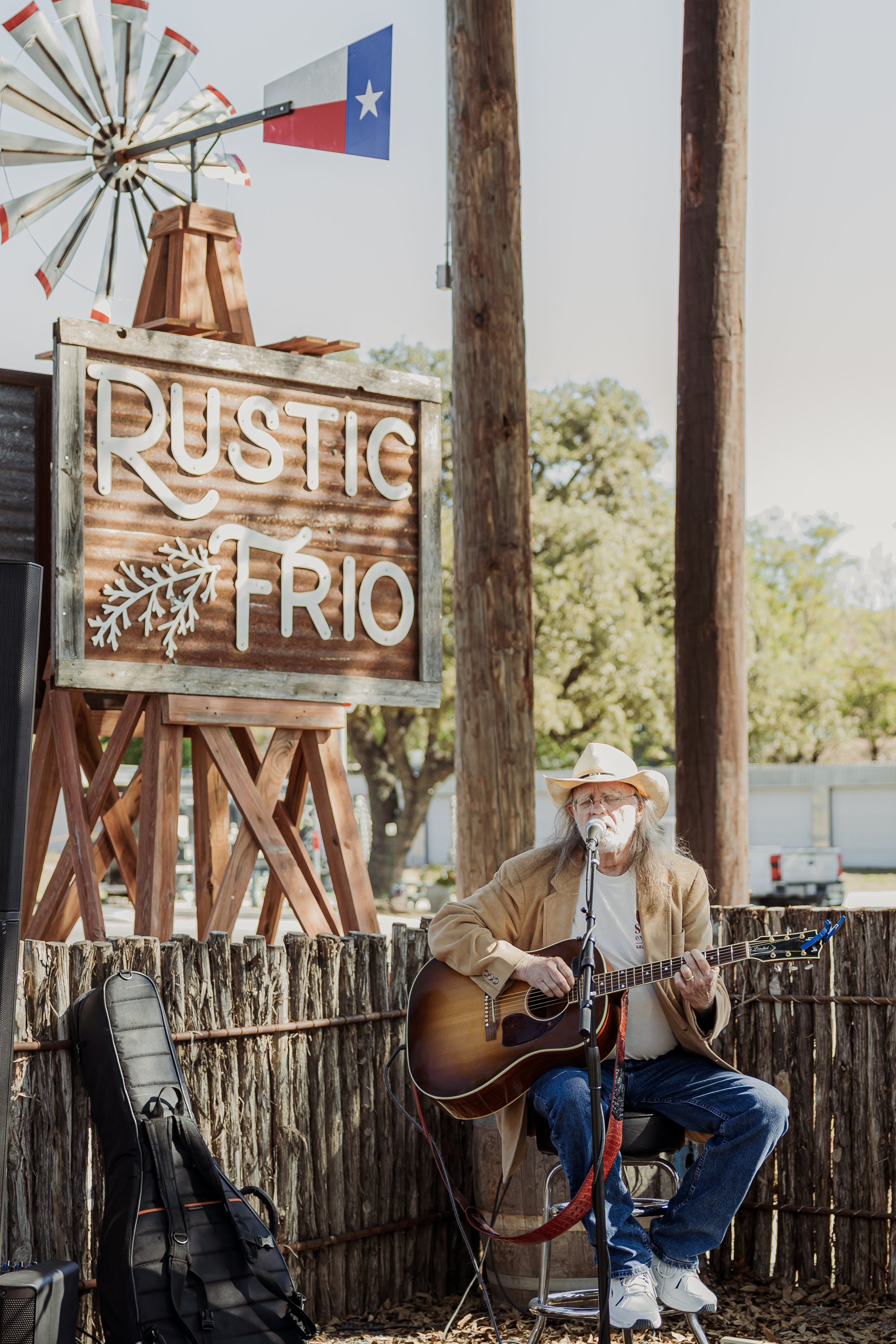 A man with long gray hair and beard, wearing a cowboy hat, playing acoustic guitar and singing into a microphone outdoors at Rustic Frio, with a large wooden sign, trees, and a windmill in the background.