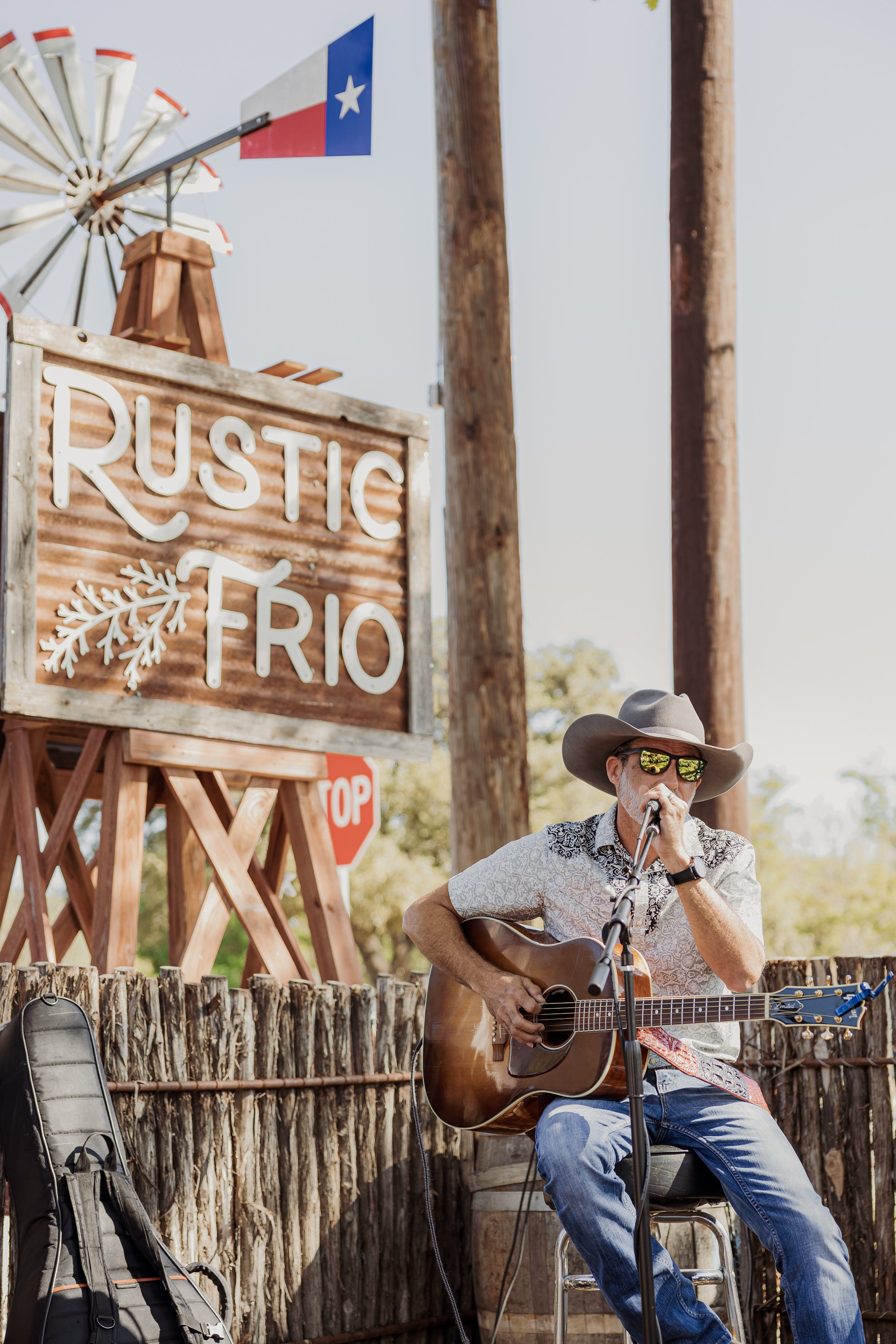 Musician wearing a cowboy hat and sunglasses playing guitar and singing into a microphone outdoors in front of a sign that says 'Rustic Rio' and a Texas flag.
