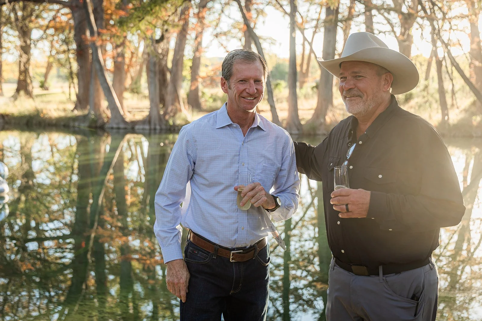 Two men stand outdoors near a body of water, holding glasses with drinks, smiling, with trees in the background, during what appears to be late afternoon or early evening.