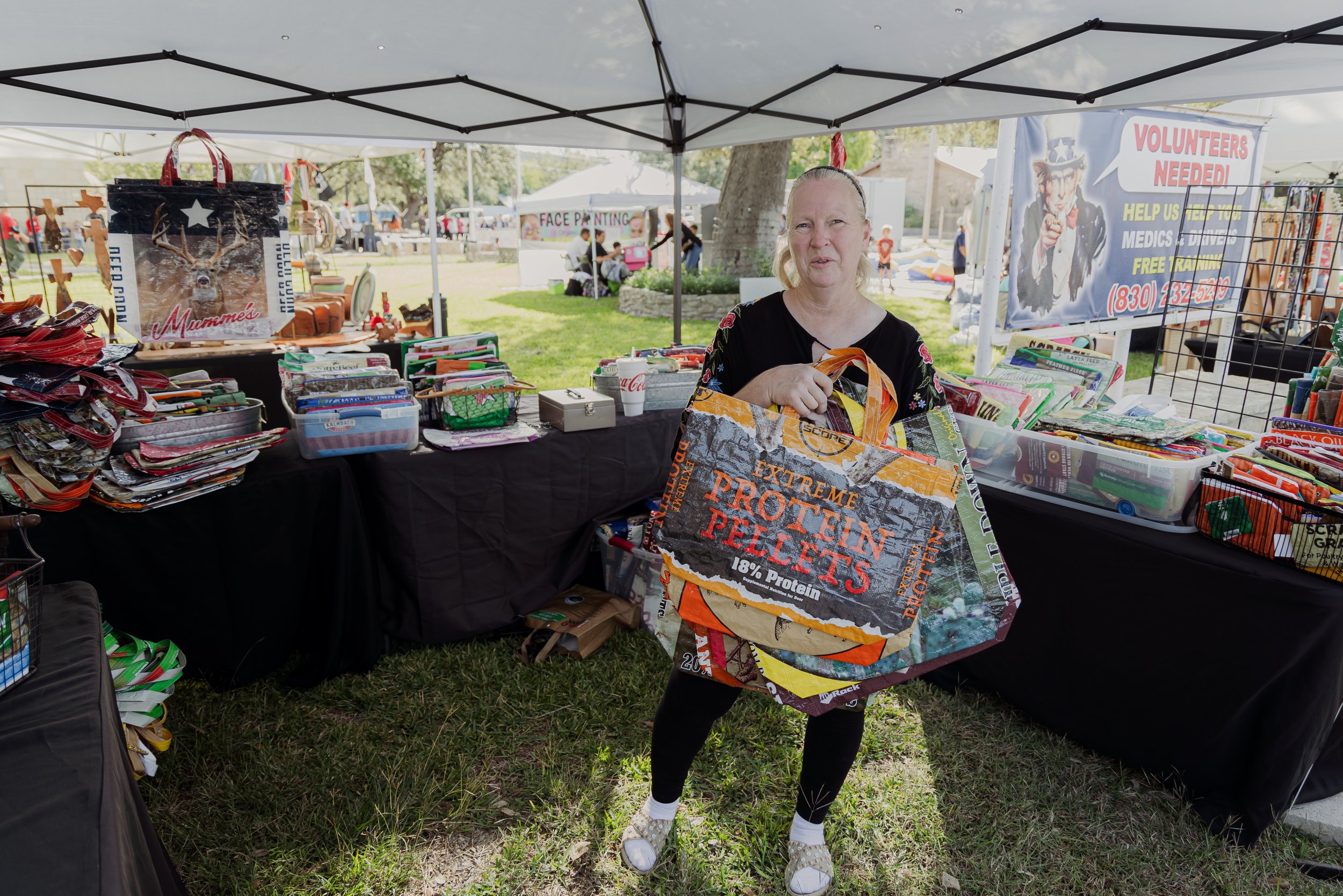 Woman standing behind a booth at an outdoor market, holding a large reusable shopping bag made of advertising banners, surrounded by tables with various items for sale, with tents and people in the background.