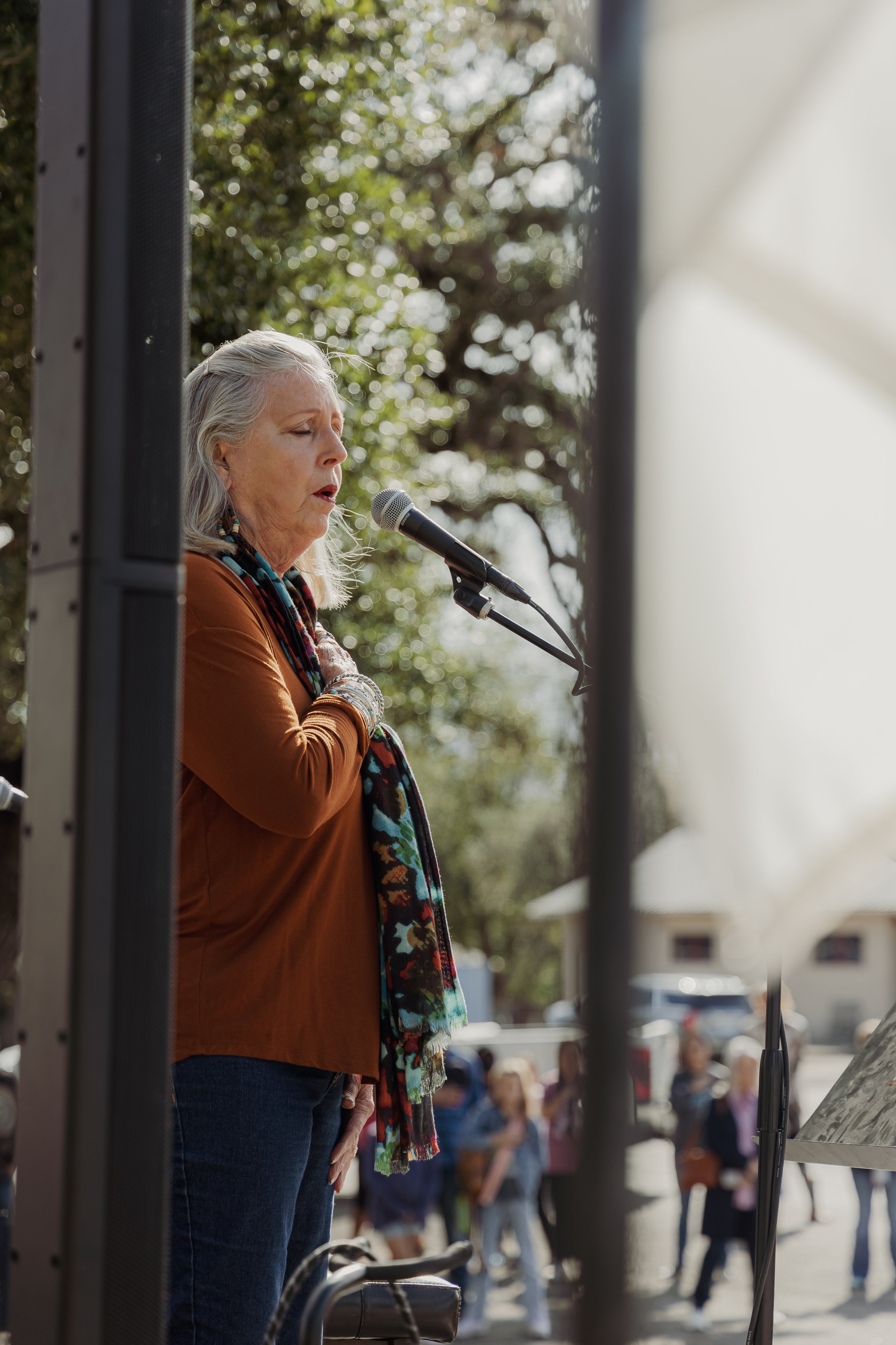 An older woman with gray hair singing into a microphone outdoors during the daytime, with a group of people and trees in the background.
