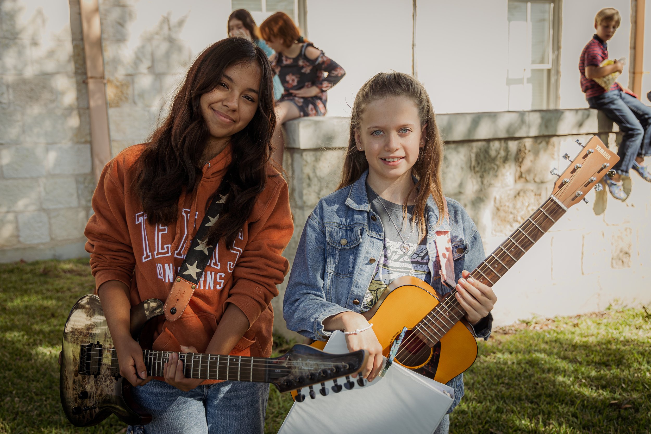 Two young girls standing outside, holding guitars, smiling at the camera with a brick wall and other children in the background.
