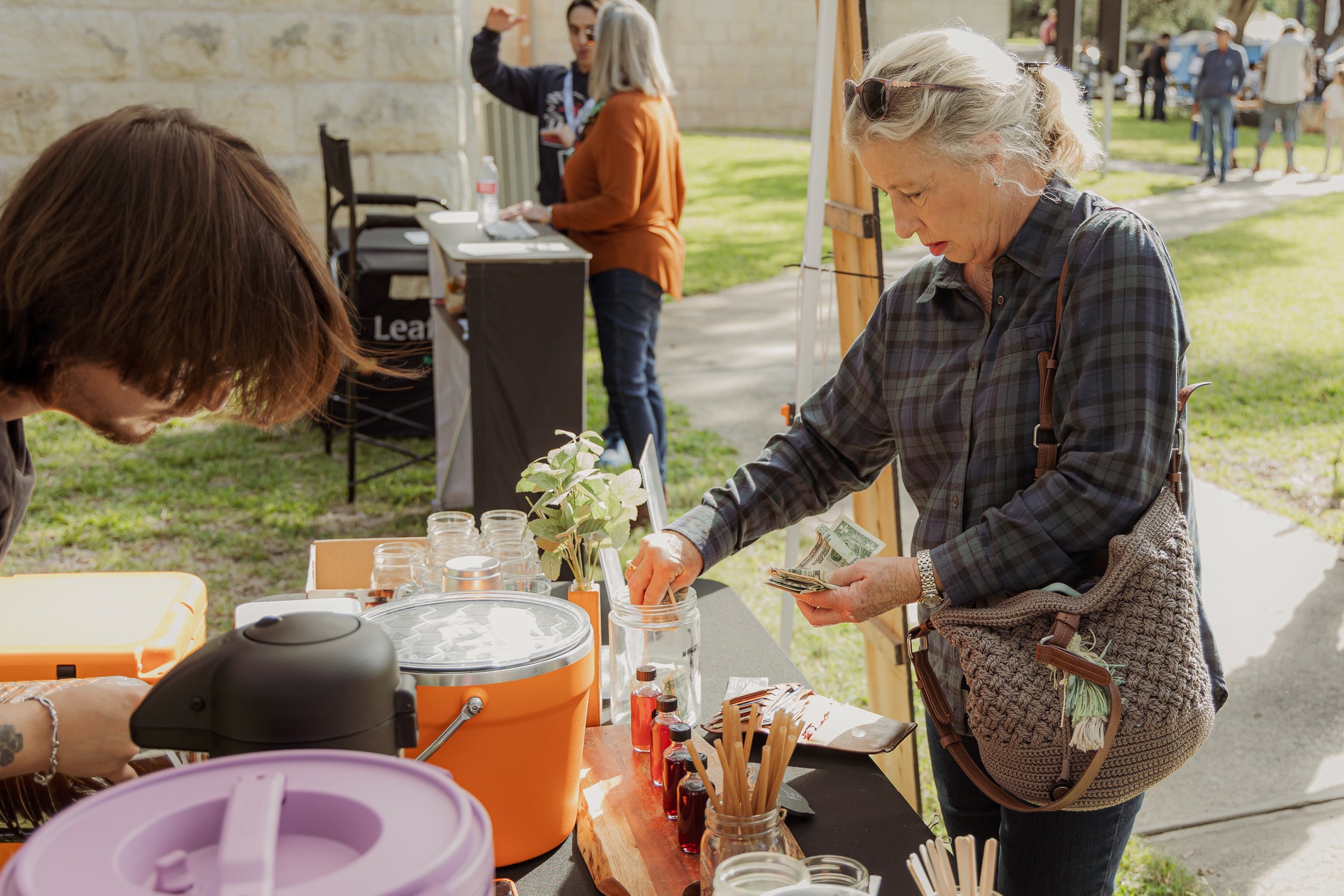 A woman is at an outdoor market booth, paying cash for a jar of product, with various small bottles and jars on the table. Other people are visible in the background.