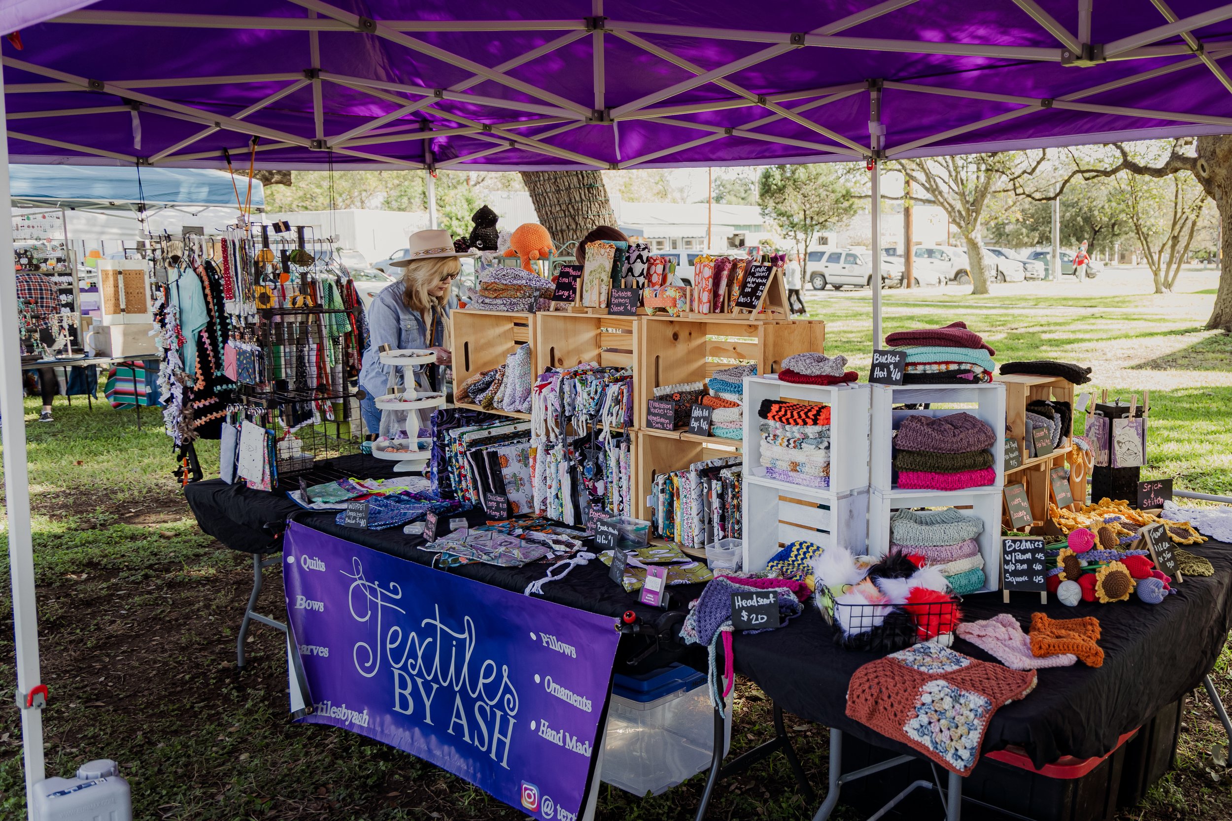 Outdoor craft fair booth selling handmade textiles, hats, and accessories, with a purple canopy and a woman browsing the items.