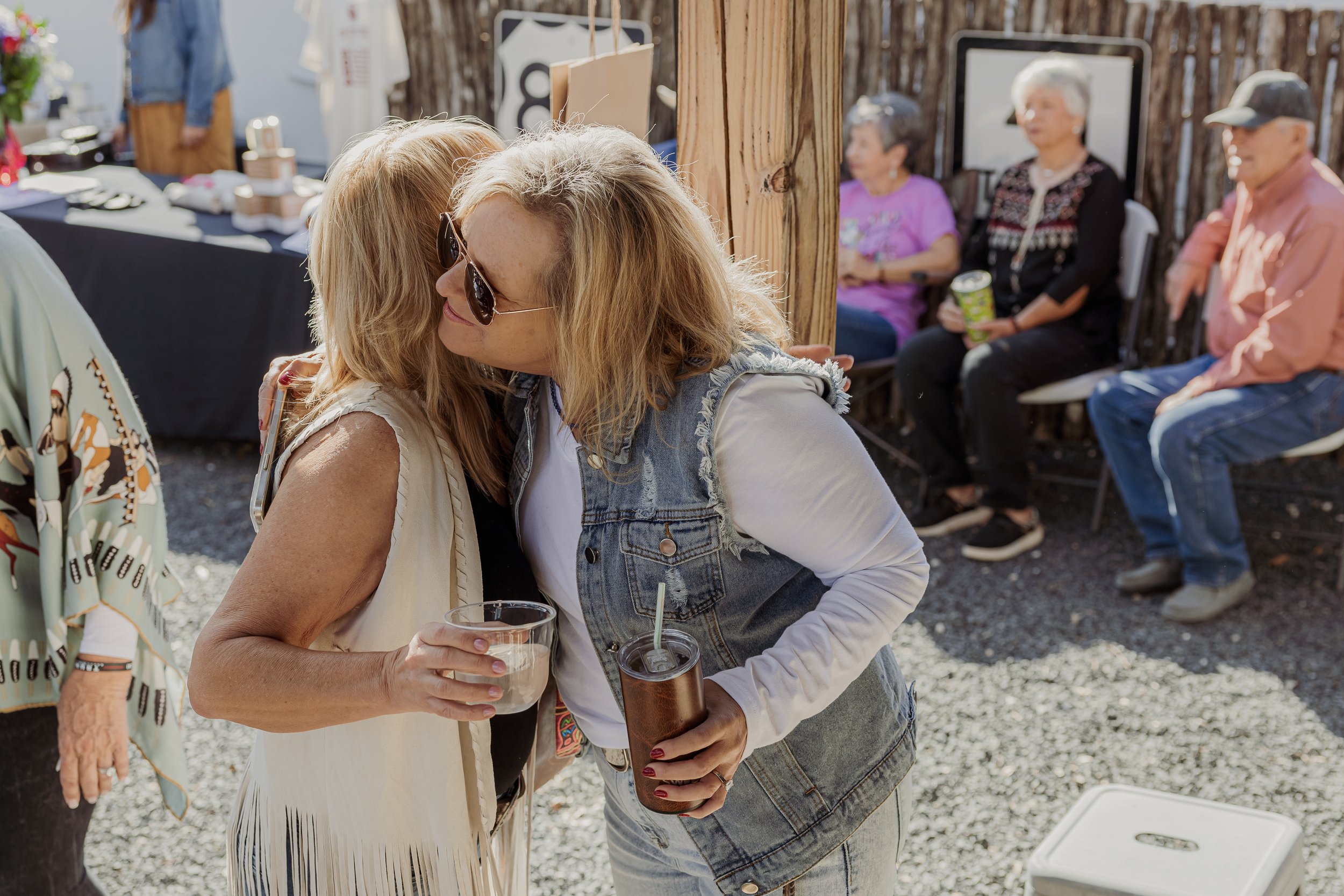 Two women hugging at an outdoor gathering, with other seated people in the background.