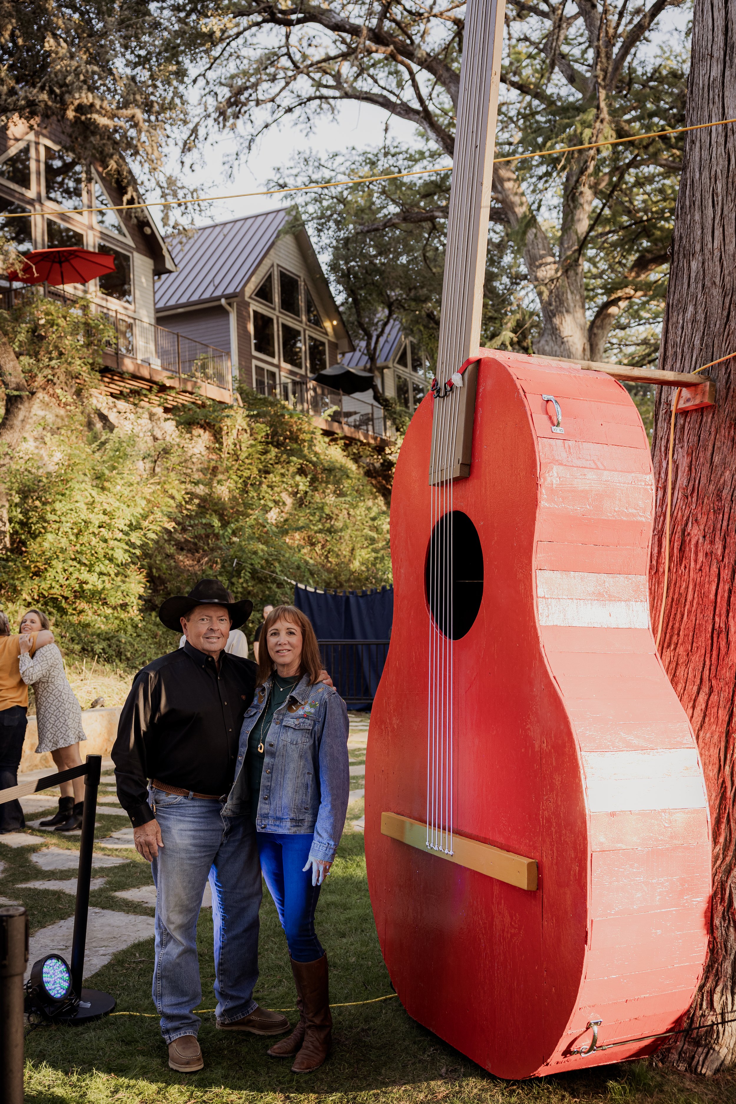 A man and woman standing next to a giant red guitar sculpture outdoors, with a hillside house and trees in the background.