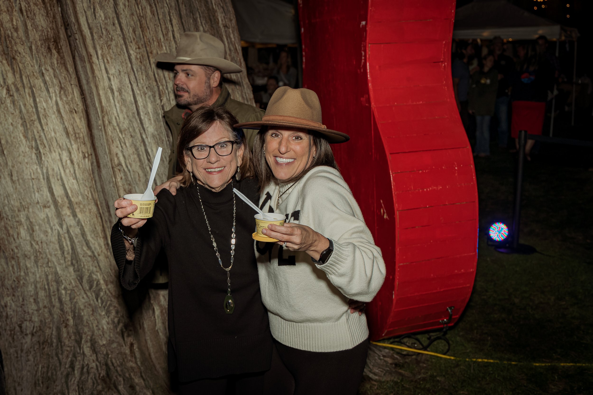 Two women smiling and holding cups of ice cream, standing in front of a large tree and a red wooden structure at an outdoor event during nighttime.