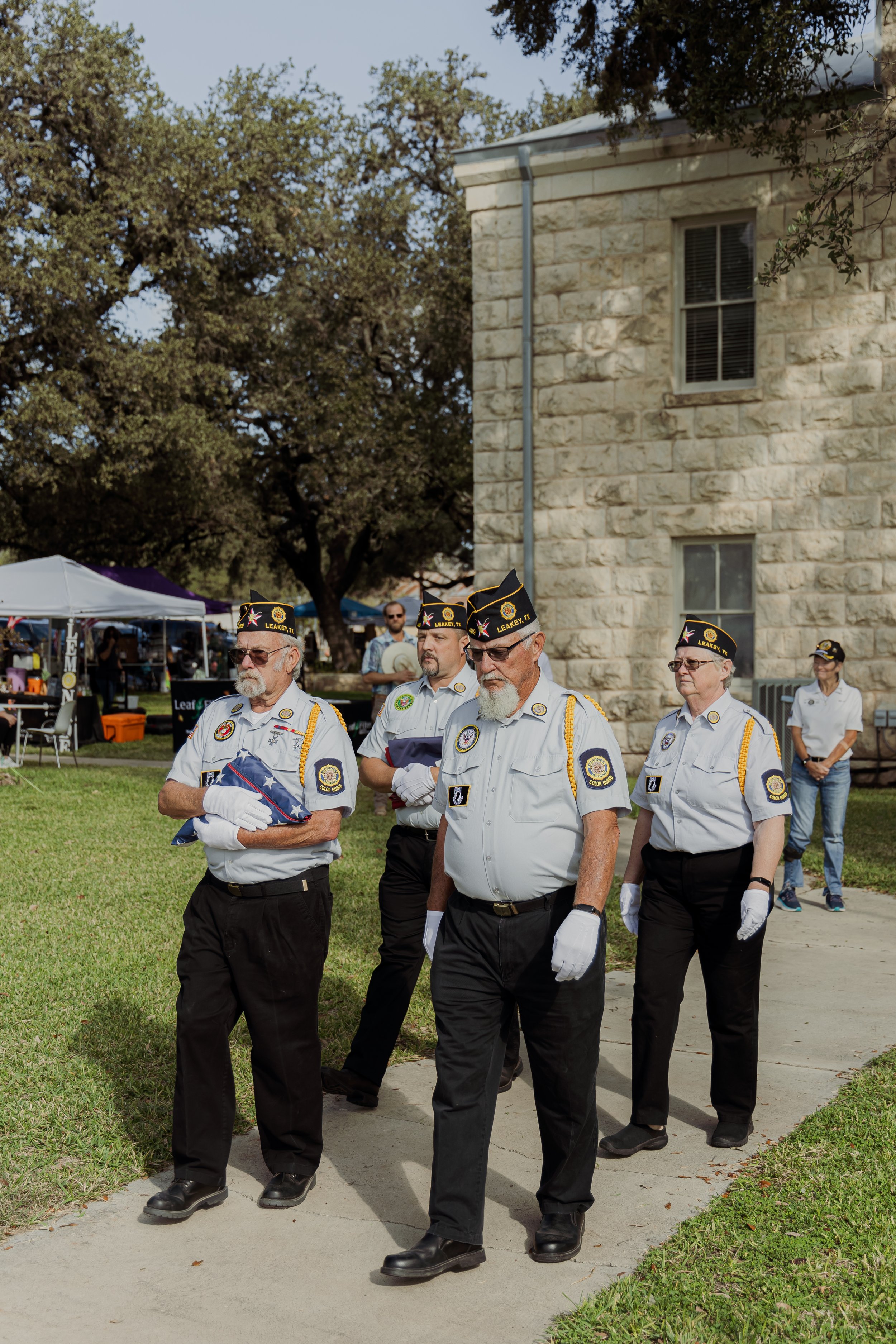 Group of elderly veterans wearing white shirts and black pants, some with black hats, participating in memorial ceremony outdoors near a stone building and trees.