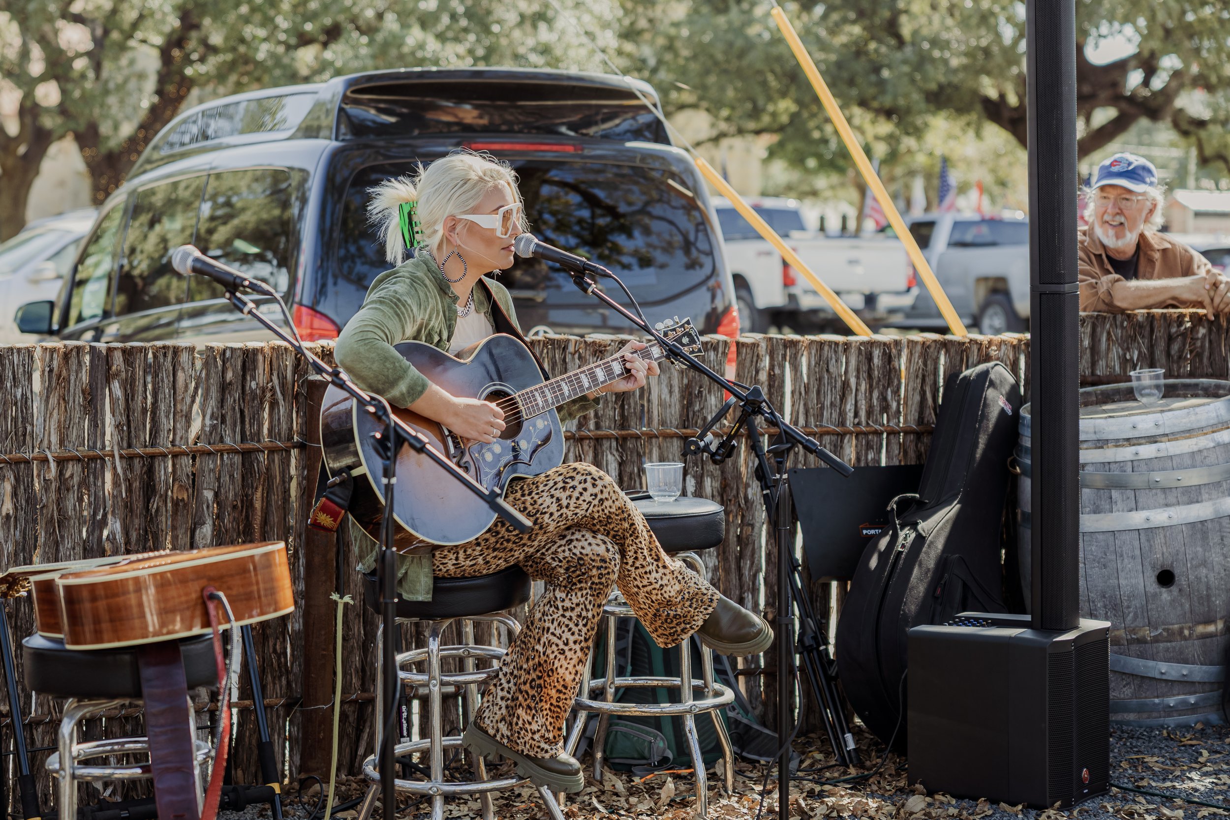 Female musician with bleached hair, sunglasses, green jacket, and animal print pants playing an acoustic guitar and singing into a microphone outdoors, with a man watching nearby.