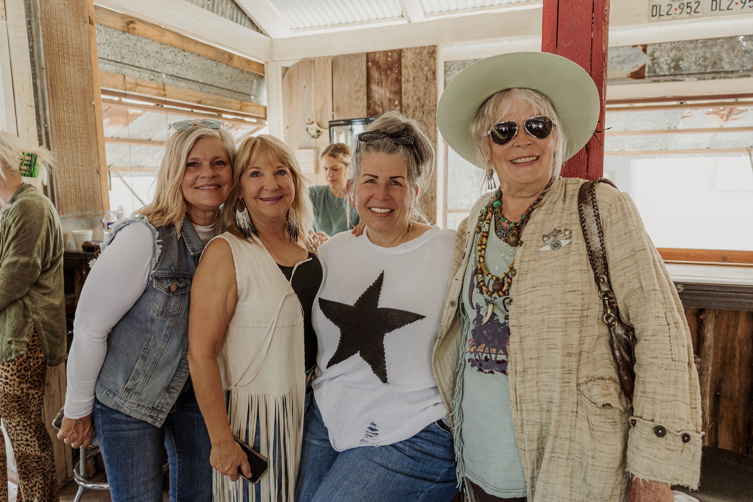 Four women smiling and posing together indoors at a social gathering, wearing casual and stylish clothing with one woman wearing sunglasses and a wide-brimmed hat.