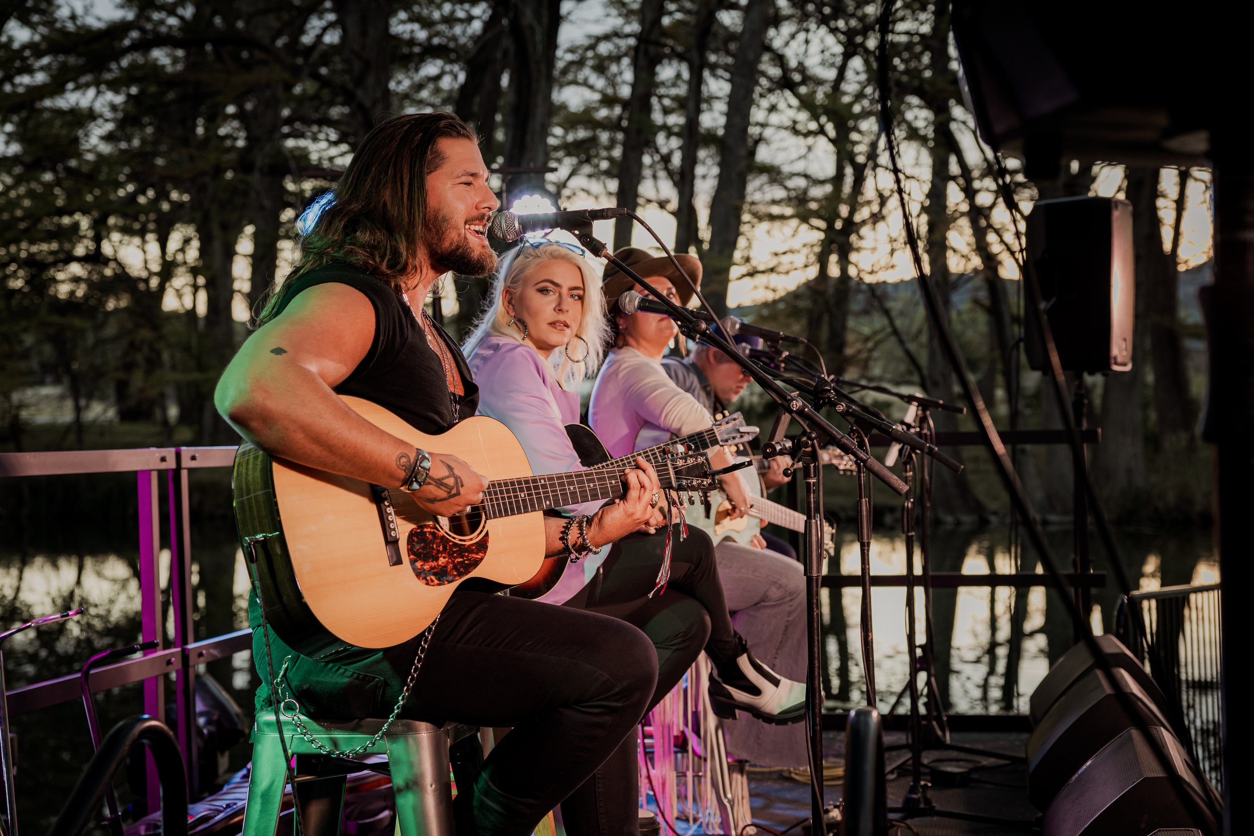 Four musicians playing guitars and singing outdoors during a sunset, with trees in the background.