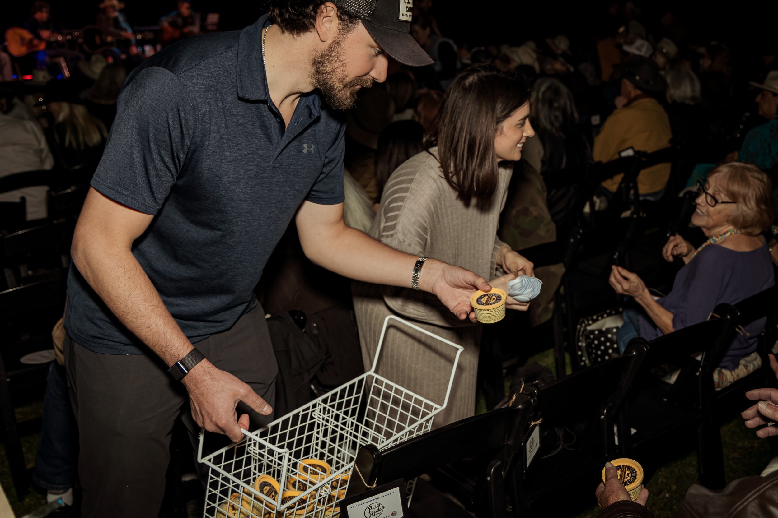 A man in a dark blue shirt and a woman in a light-colored sweater standing in front of seated audience members at an event. The man holds a white basket filled with small containers, and the woman is handing out a small tub of ice cream.