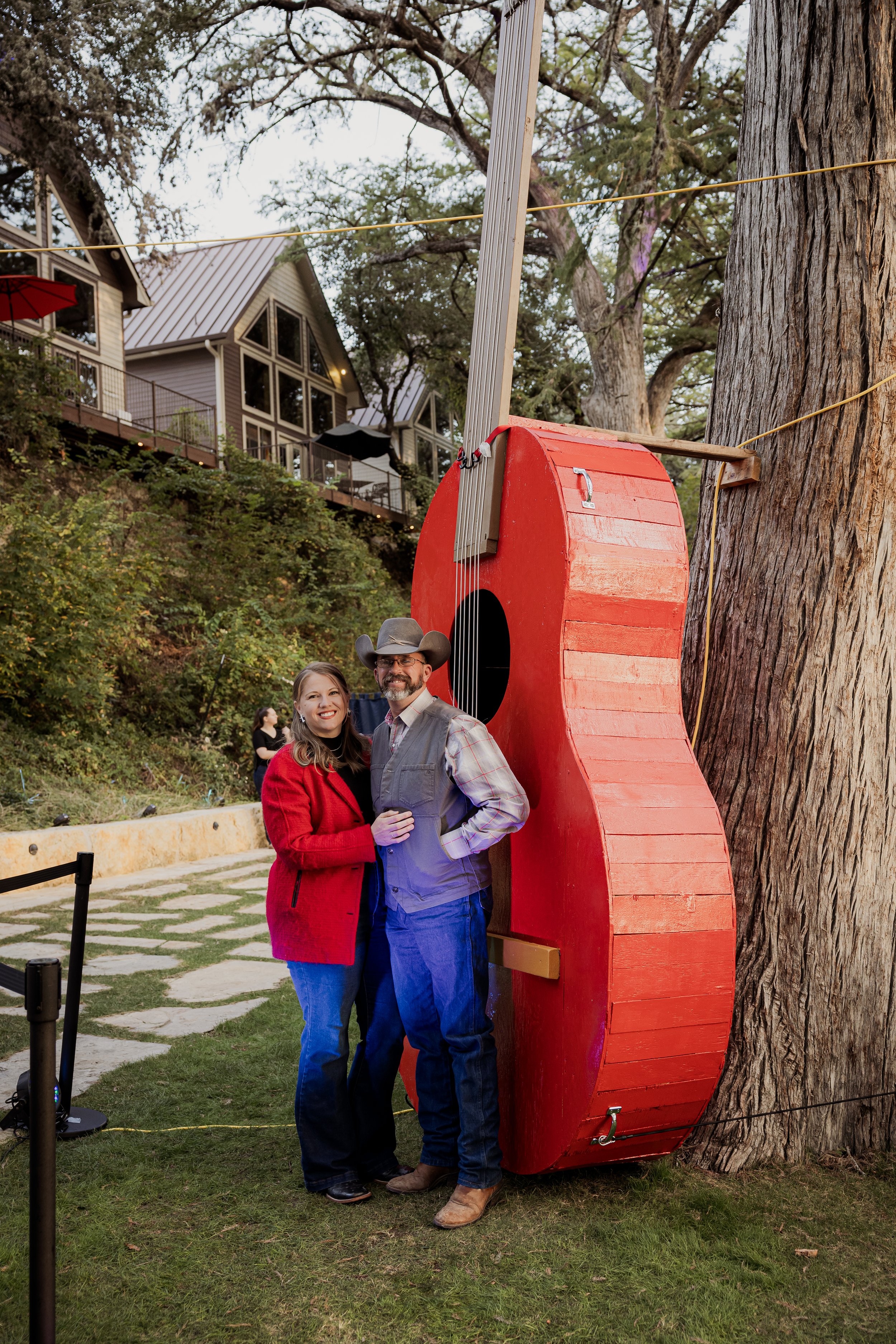 A man and woman stand together smiling in front of a large red wooden guitar sculpture attached to a tree outdoors, with houses on a hillside in the background.