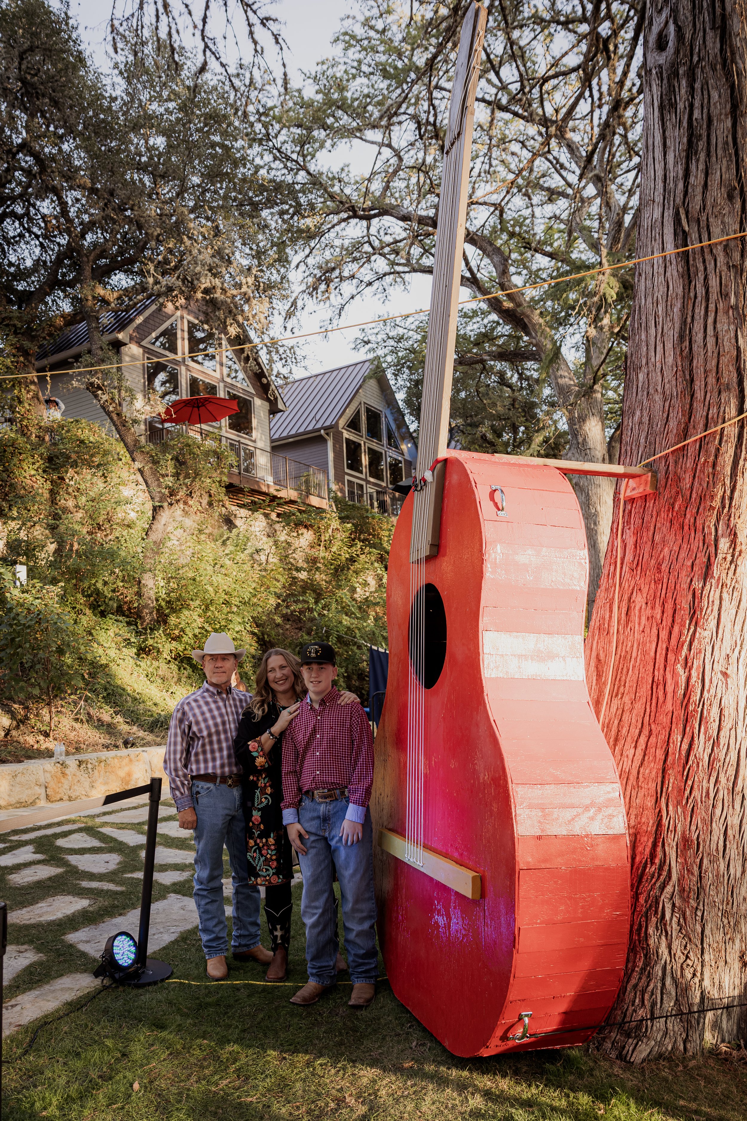 Three people standing outdoors near a large red guitar sculpture, with trees and houses in the background.