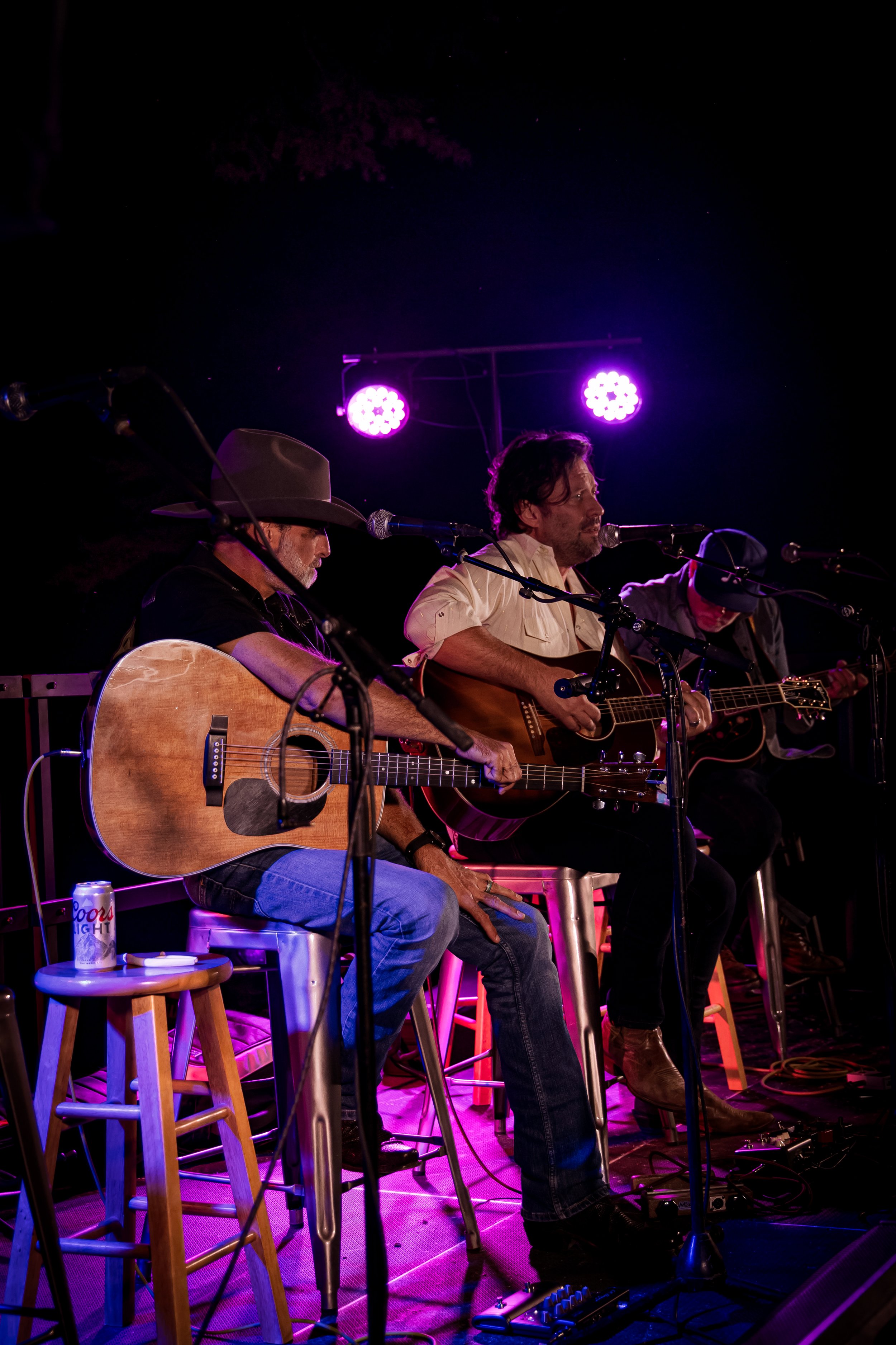 Three musicians playing acoustic guitars on stage with purple stage lights and microphones.