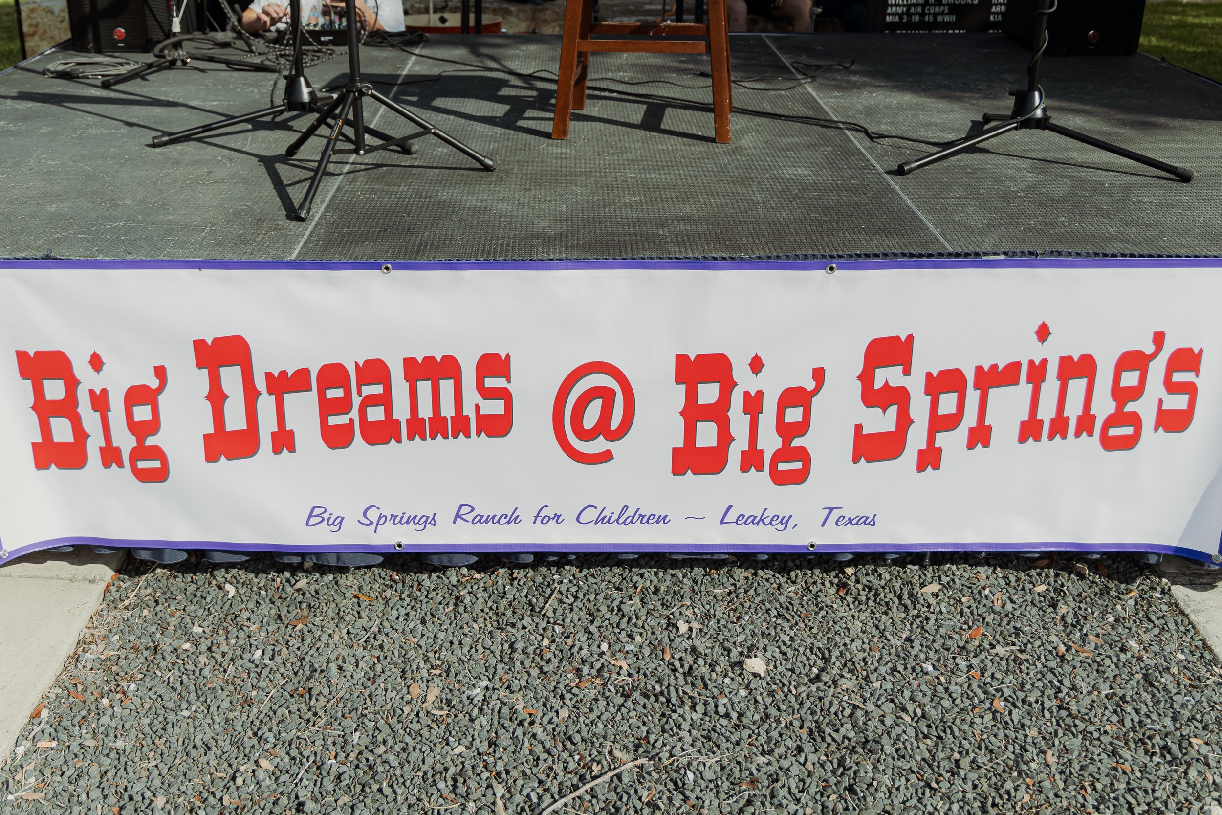 A stage with microphone stands and a wooden chair, with a large banner in front that reads 'Big Dreams @ Big Springs' in red letters, and additional text below stating 'Big Springs Ranch for Children - Leakey, Texas.'