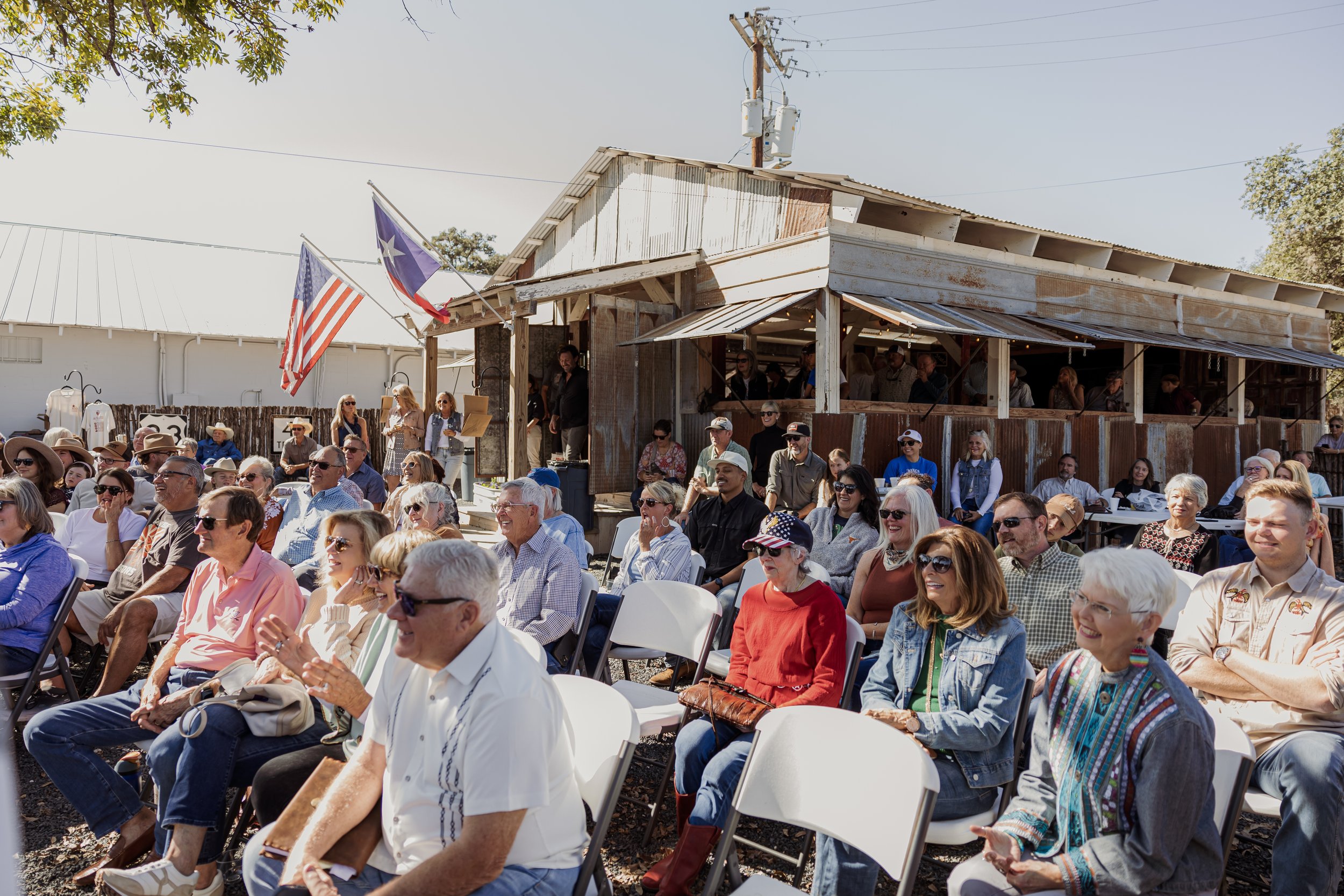 People seated outdoors at a gathering or event, with a rustic building and American flags in the background.