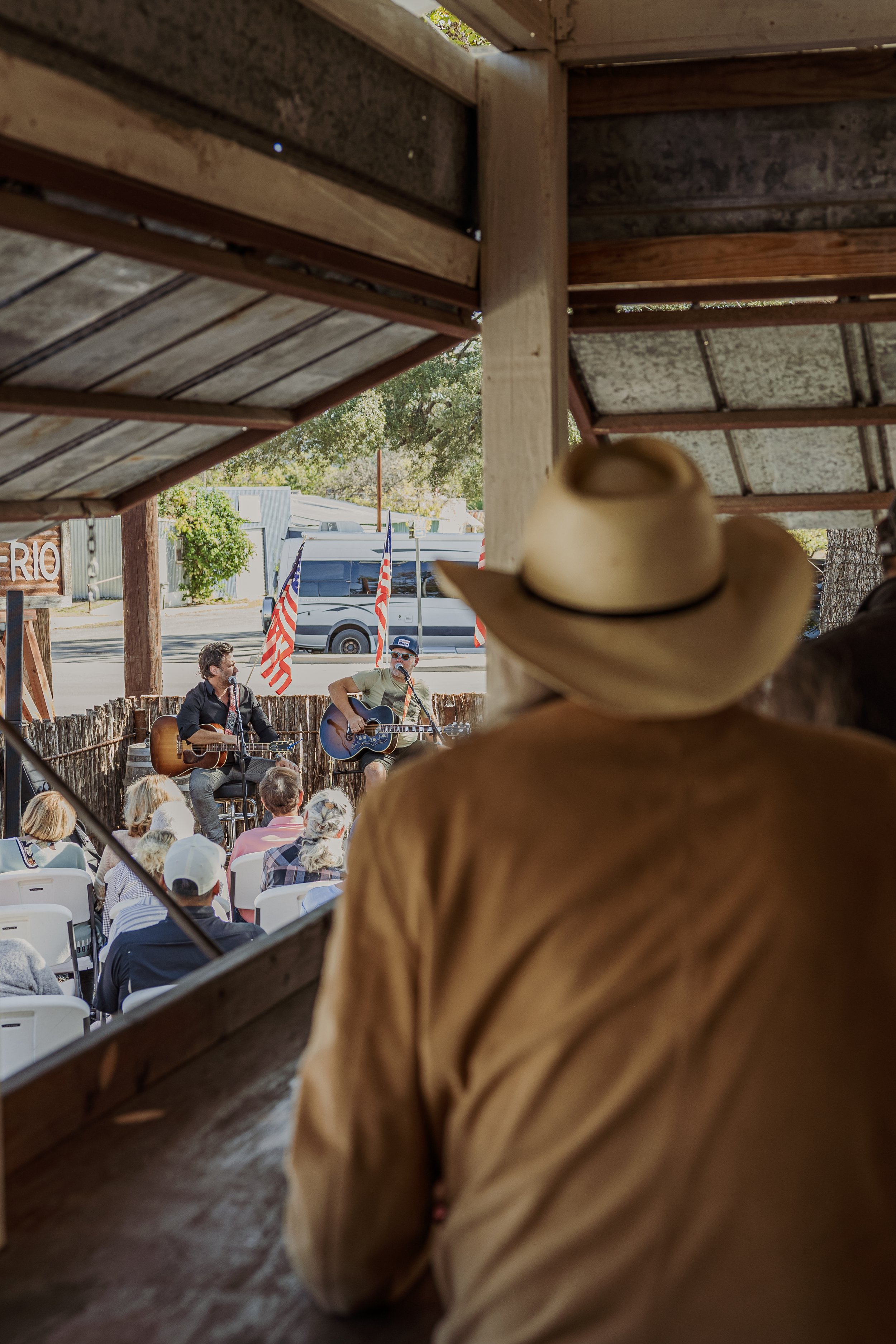 A person wearing a cowboy hat and tan shirt watching a live music performance at an outdoor venue with American flags.