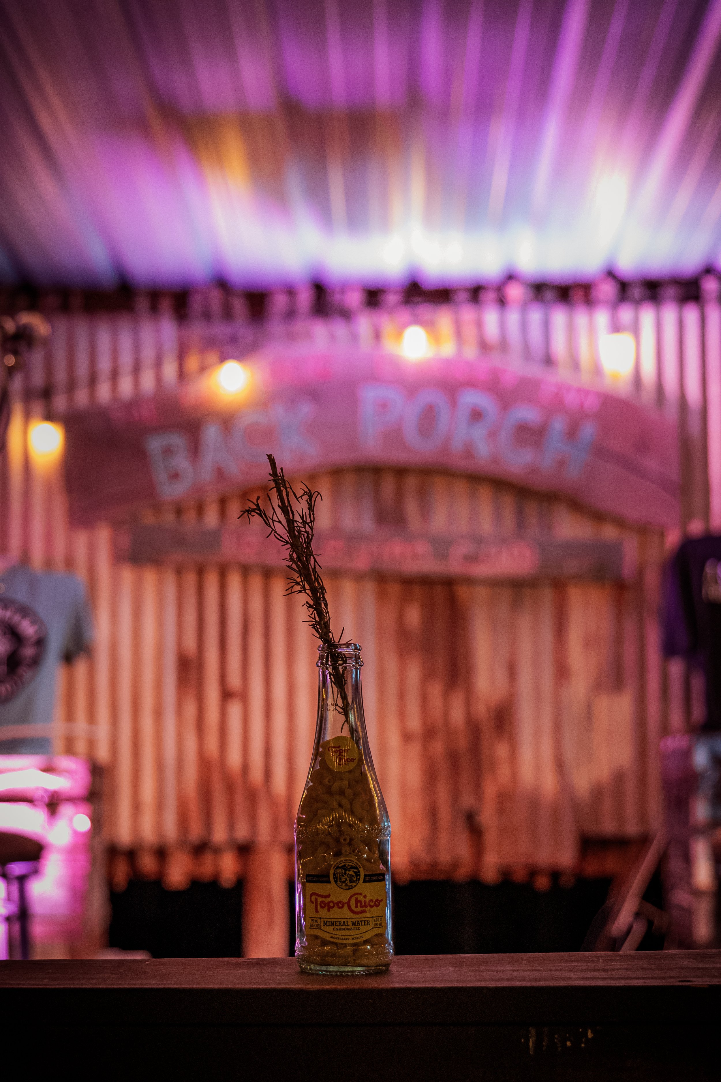 A glass bottle of Topo Chico mineral water with a dried herb sprig inside, placed on a wooden surface in a dimly lit establishment with a neon pink sign that says 'Back Porch' in the background.