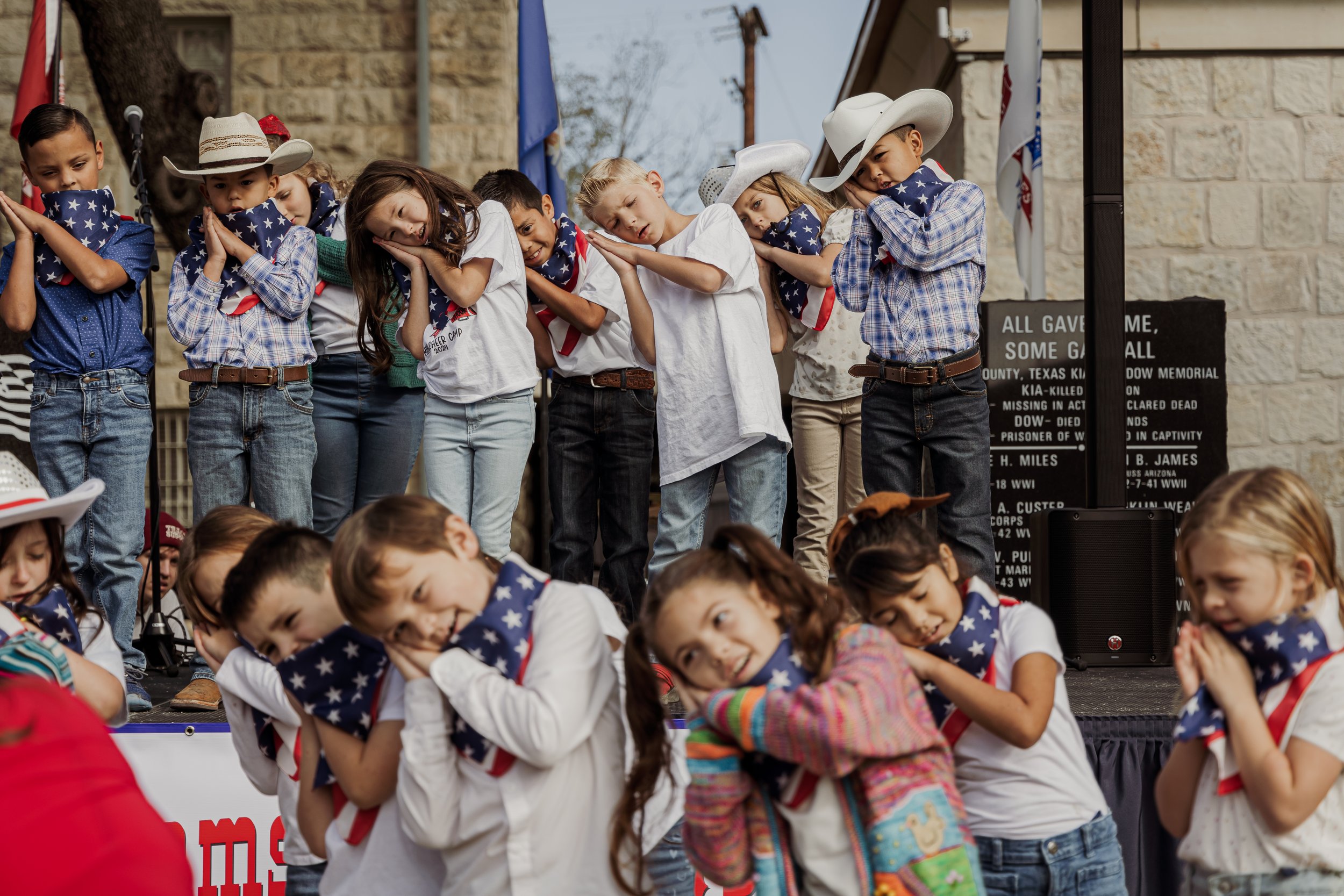 Children dressed in patriotic clothing performing a patriotic song or dance on a stage during a Memorial Day or Fourth of July event.