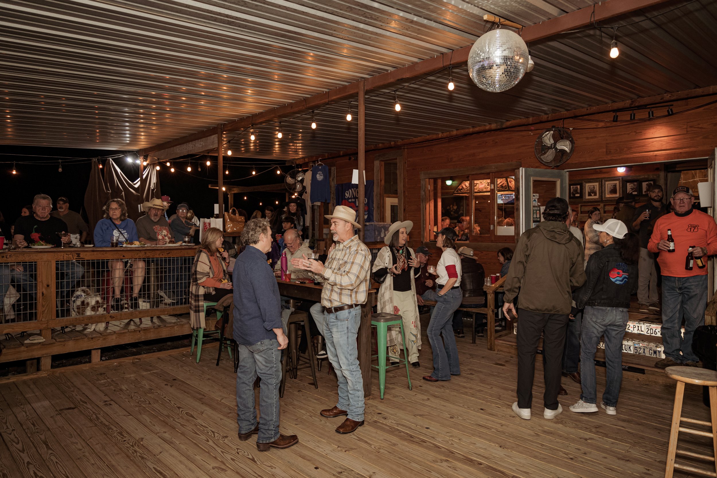 A lively indoor social gathering at a rustic bar with string lights, a disco ball, and a crowd of people talking, drinking, and enjoying the evening.