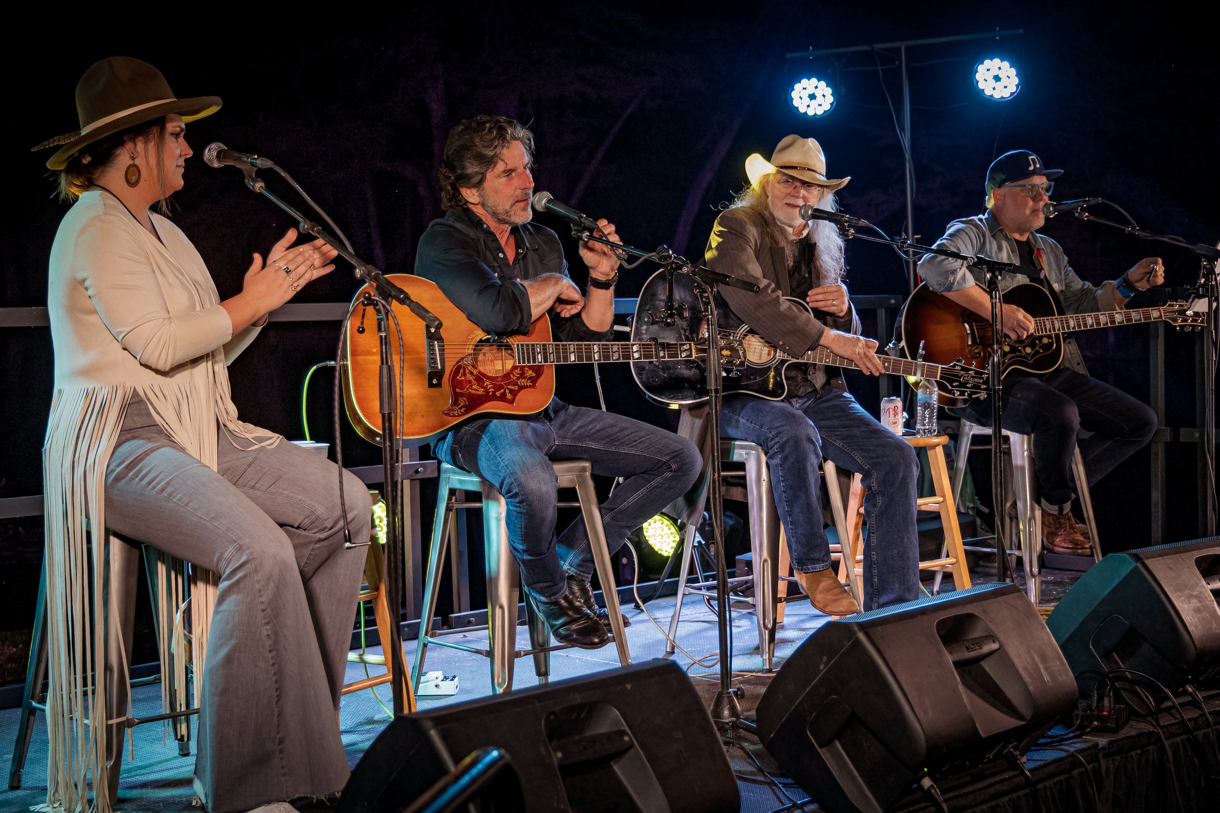 Four musicians performing on a stage at night, two men playing guitars, one woman in a cowboy hat, and a woman clapping, with stage lights behind them.