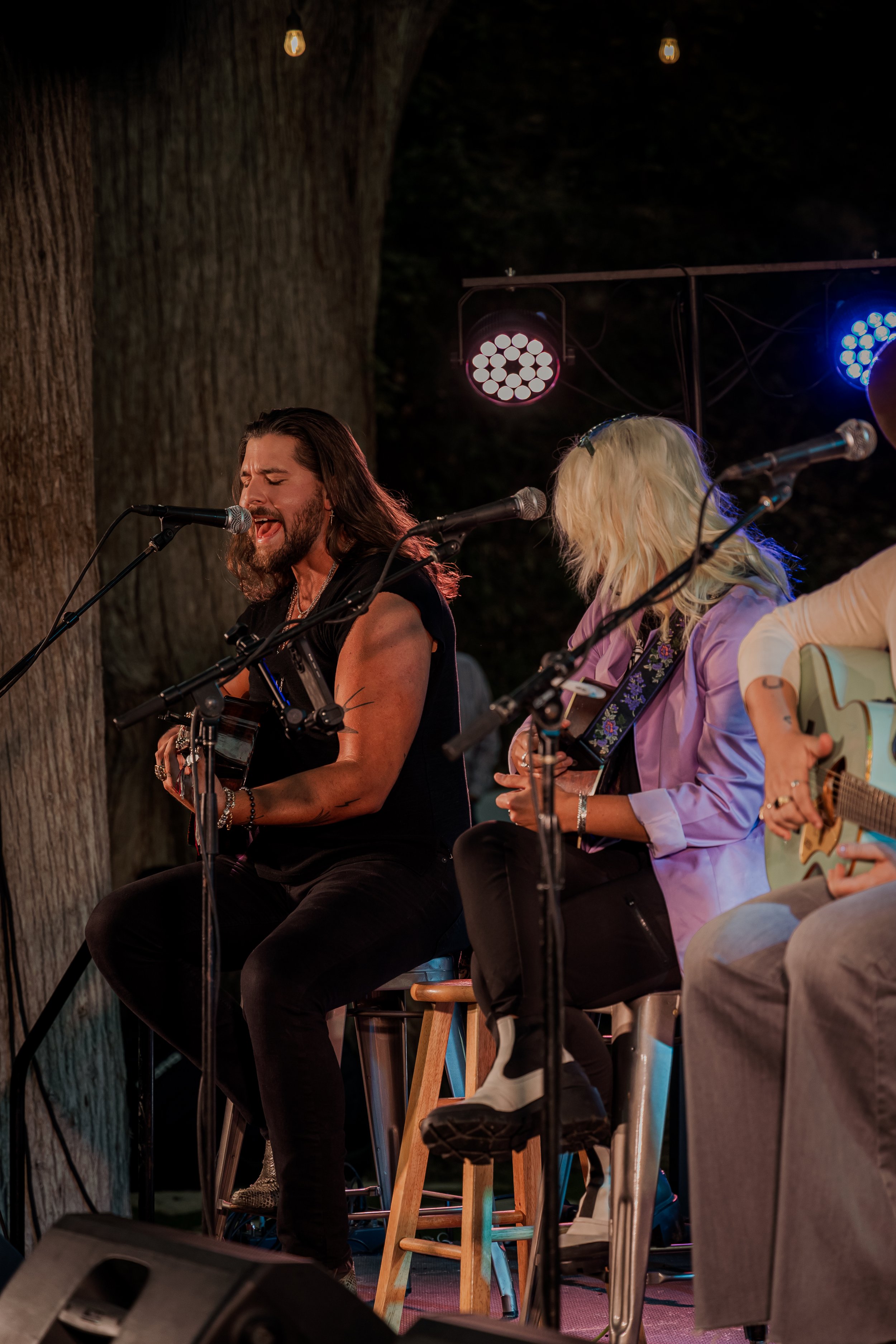 Musicians performing on stage with microphones, guitars, and stools, under string lights at an outdoor concert at night.