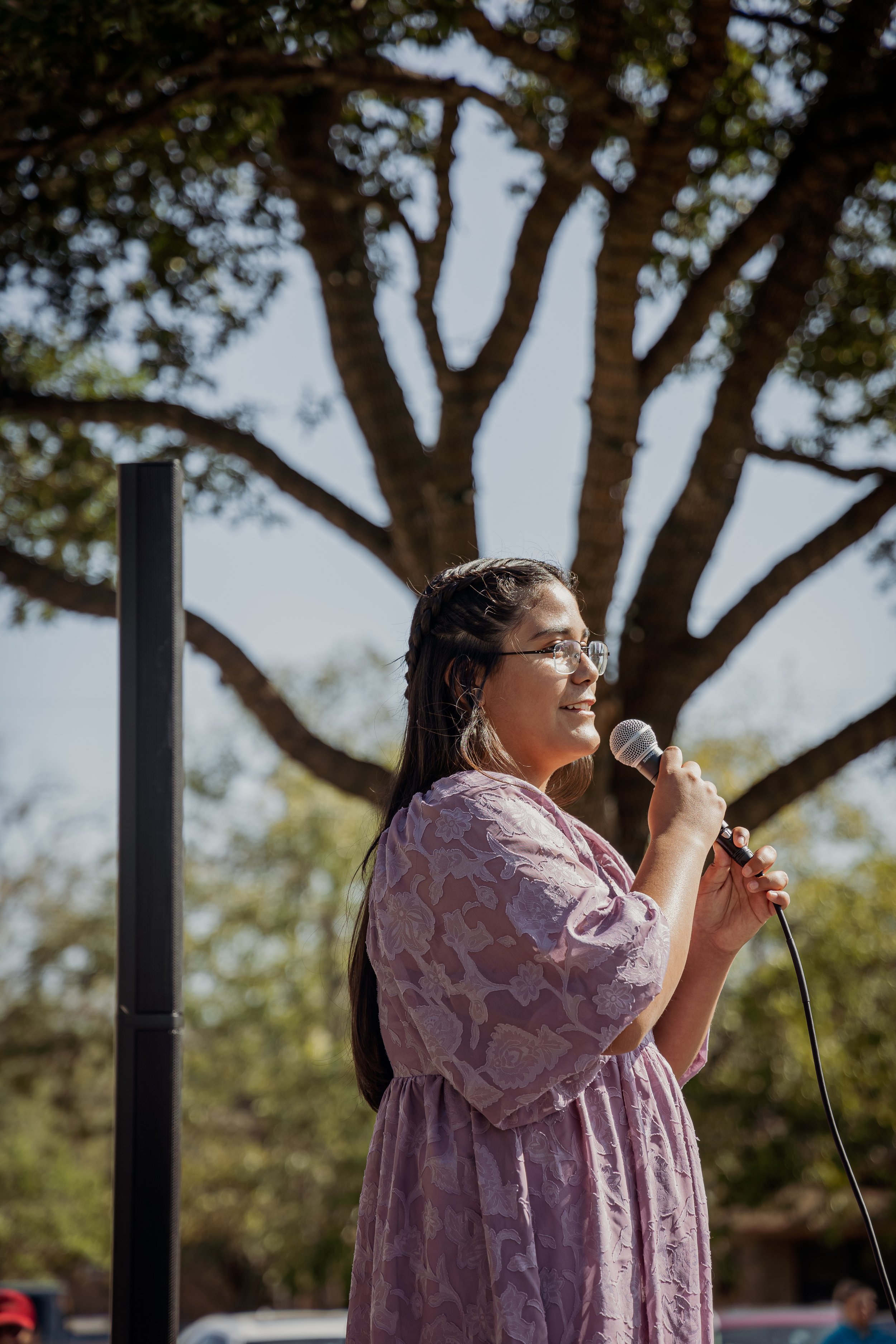 A woman with glasses and long hair wearing a pink floral dress, holding a microphone and speaking outdoors with a tree and blue sky in the background.