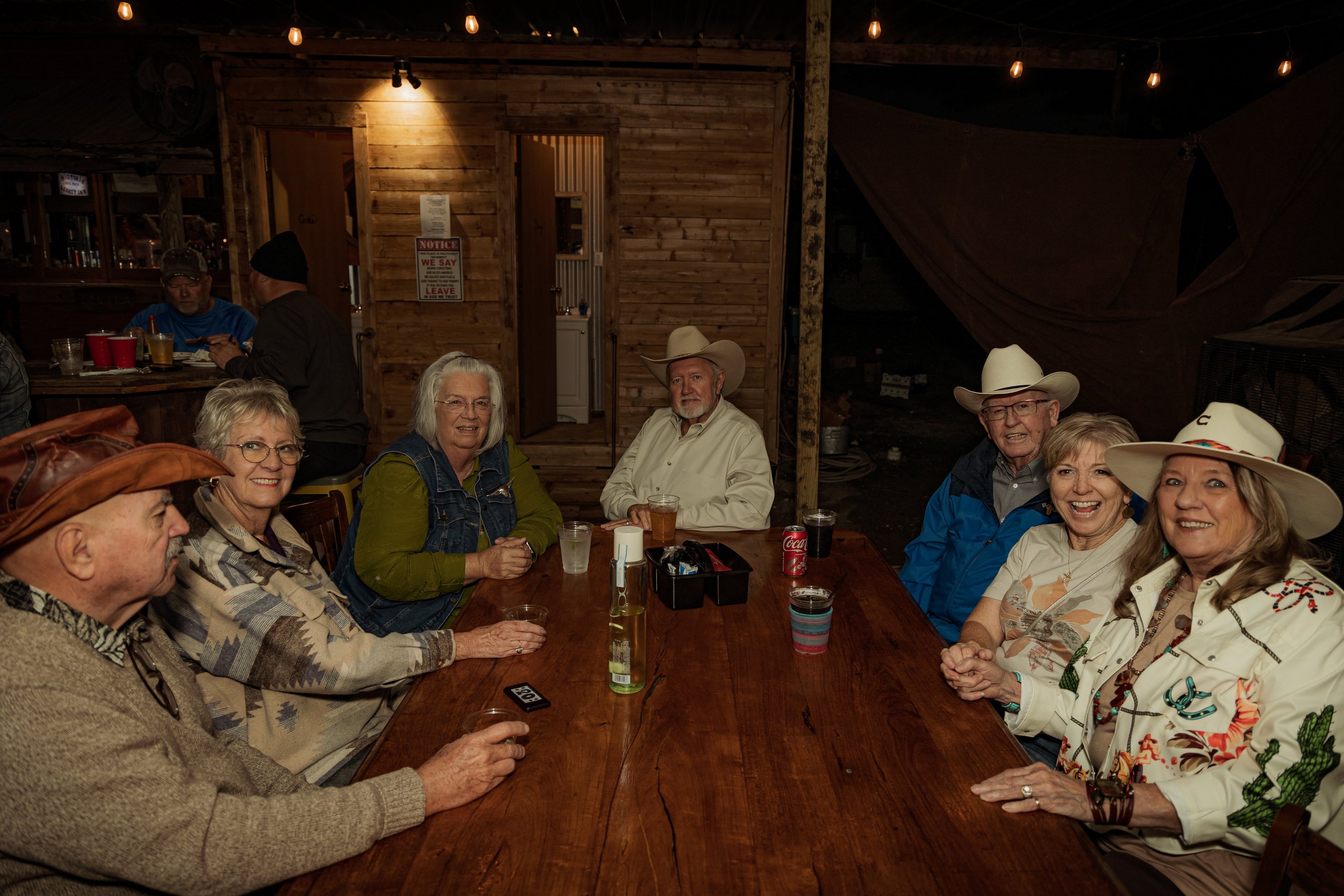 Group of seven older adults, five women and two men, sitting around a wooden table at a rustic indoor gathering, some wearing cowboy hats, with drinks and casual attire, in a warmly lit setting.