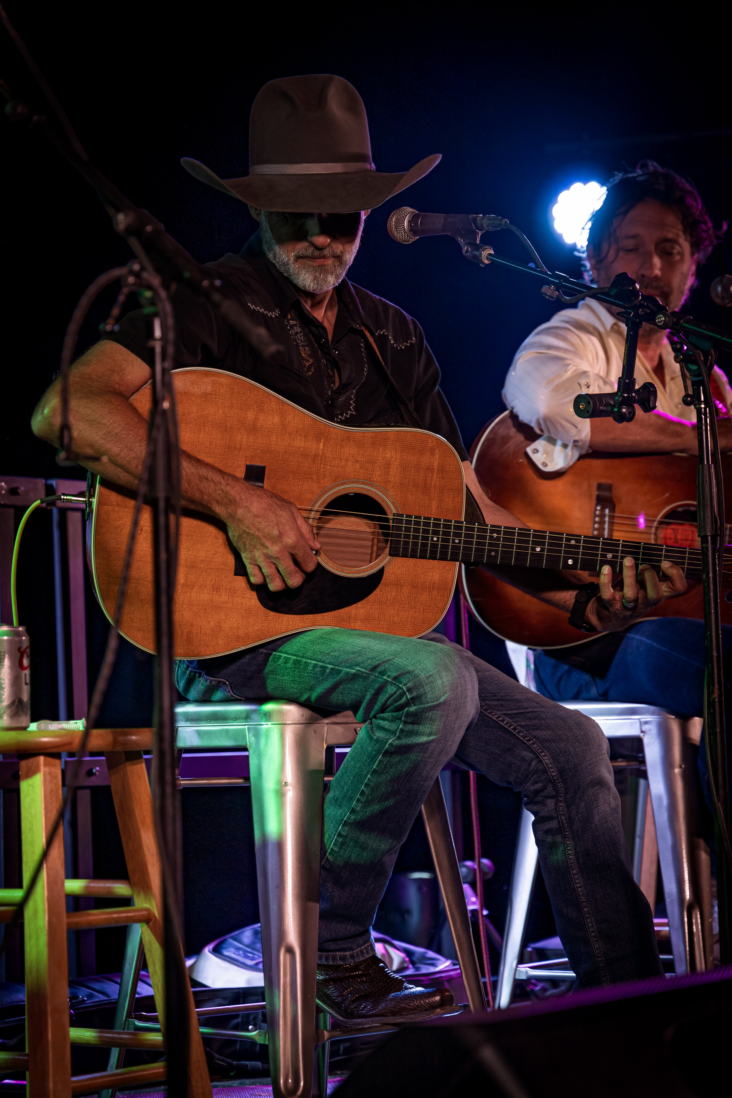 Two musicians sitting on stools playing acoustic guitars during a performance, with stage lighting in the background.