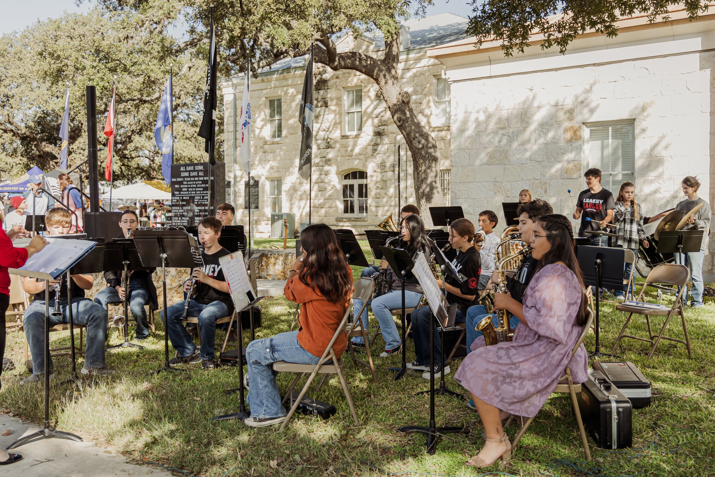 A group of children and teenagers performing in a band outdoors during daytime, with musical instruments including clarinets and saxophones, set against a backdrop of a white building, flags, and trees.