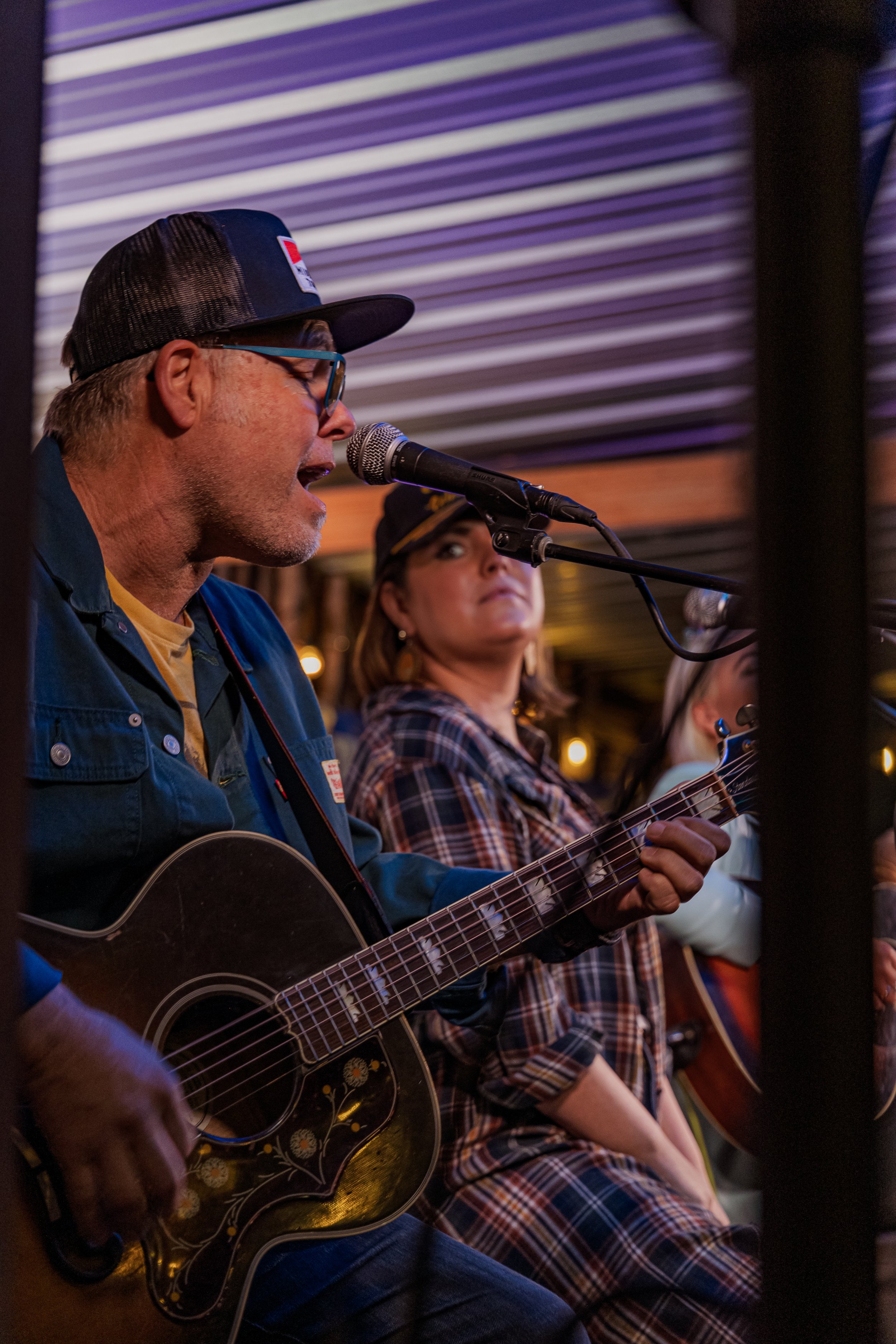 A man with glasses and a trucker hat singing into a microphone while playing an acoustic guitar, with a woman in plaid shirt in the background listening.