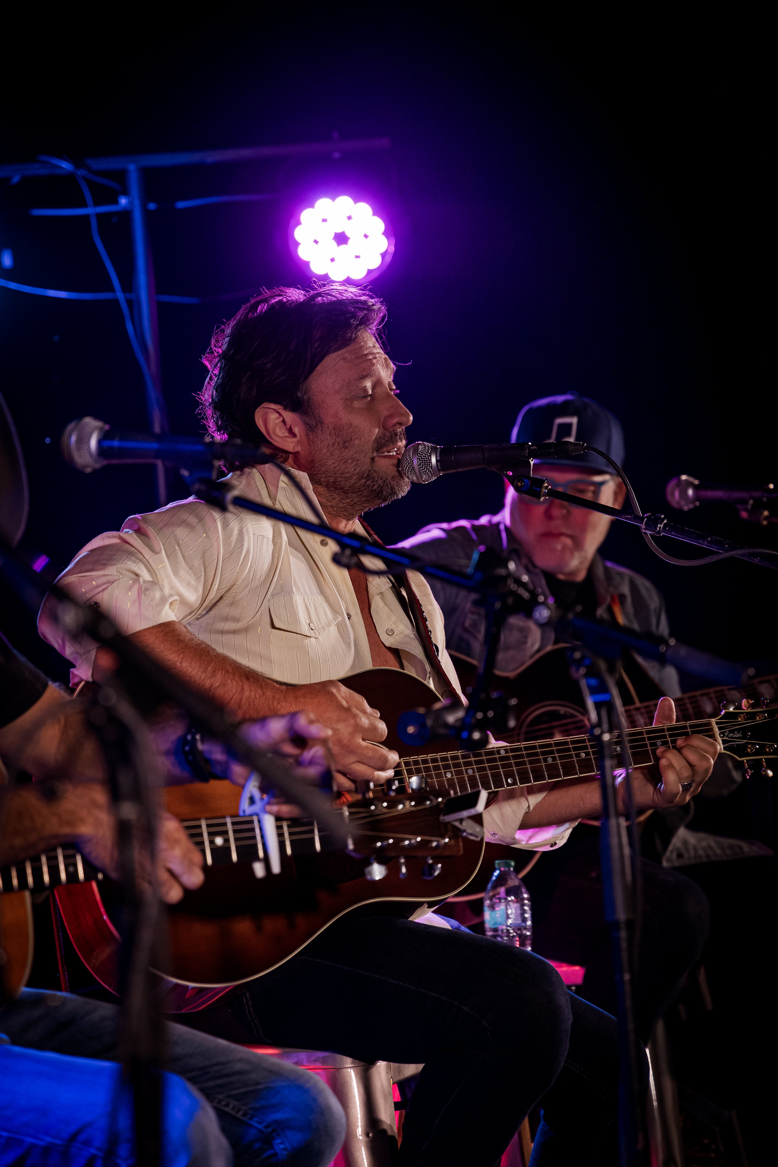 Musicians performing live on stage, singing and playing acoustic guitars under purple stage lighting, with microphones and equipment visible.