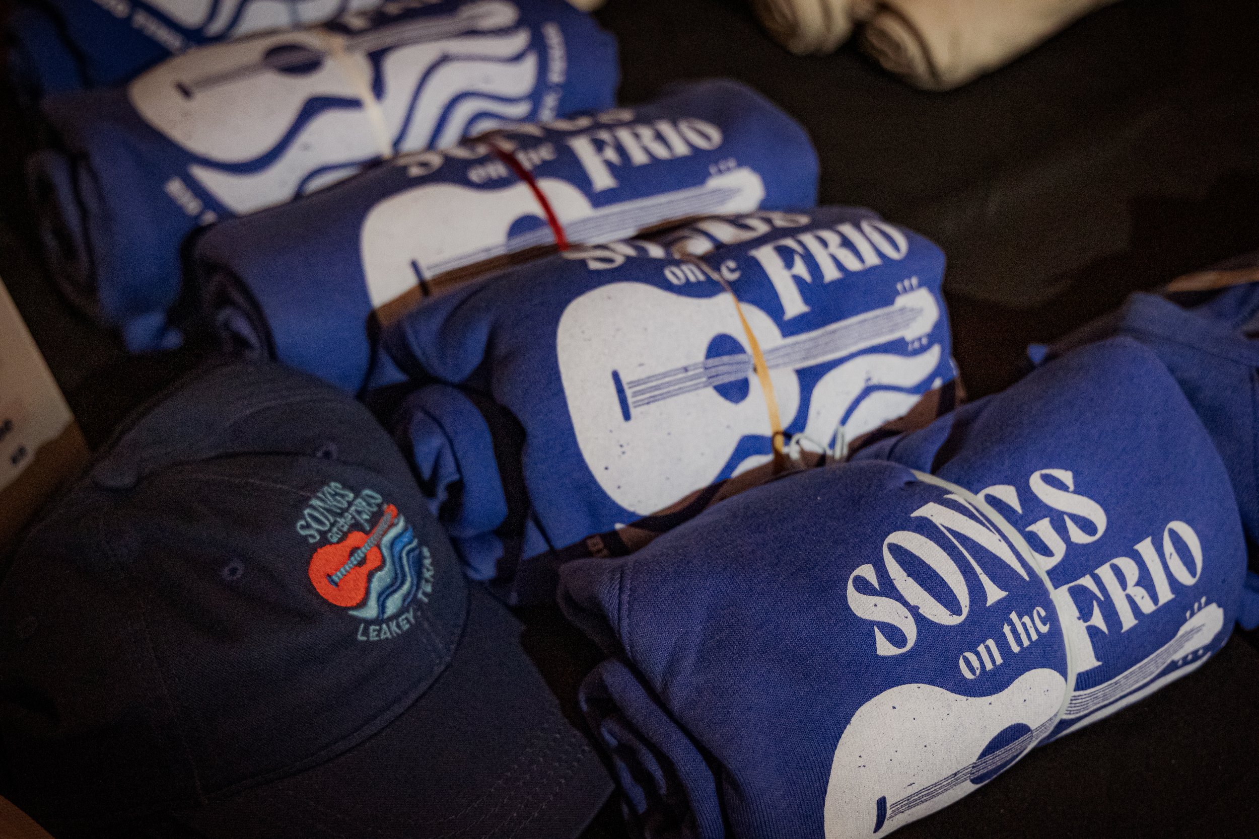 Rolled-up banners with the text "Sounds on the Frio" and a guitar graphic, a black baseball cap with a red guitar logo and the words "Sounds on the Frio", and other rolled-up cloths or banners on a dark surface.