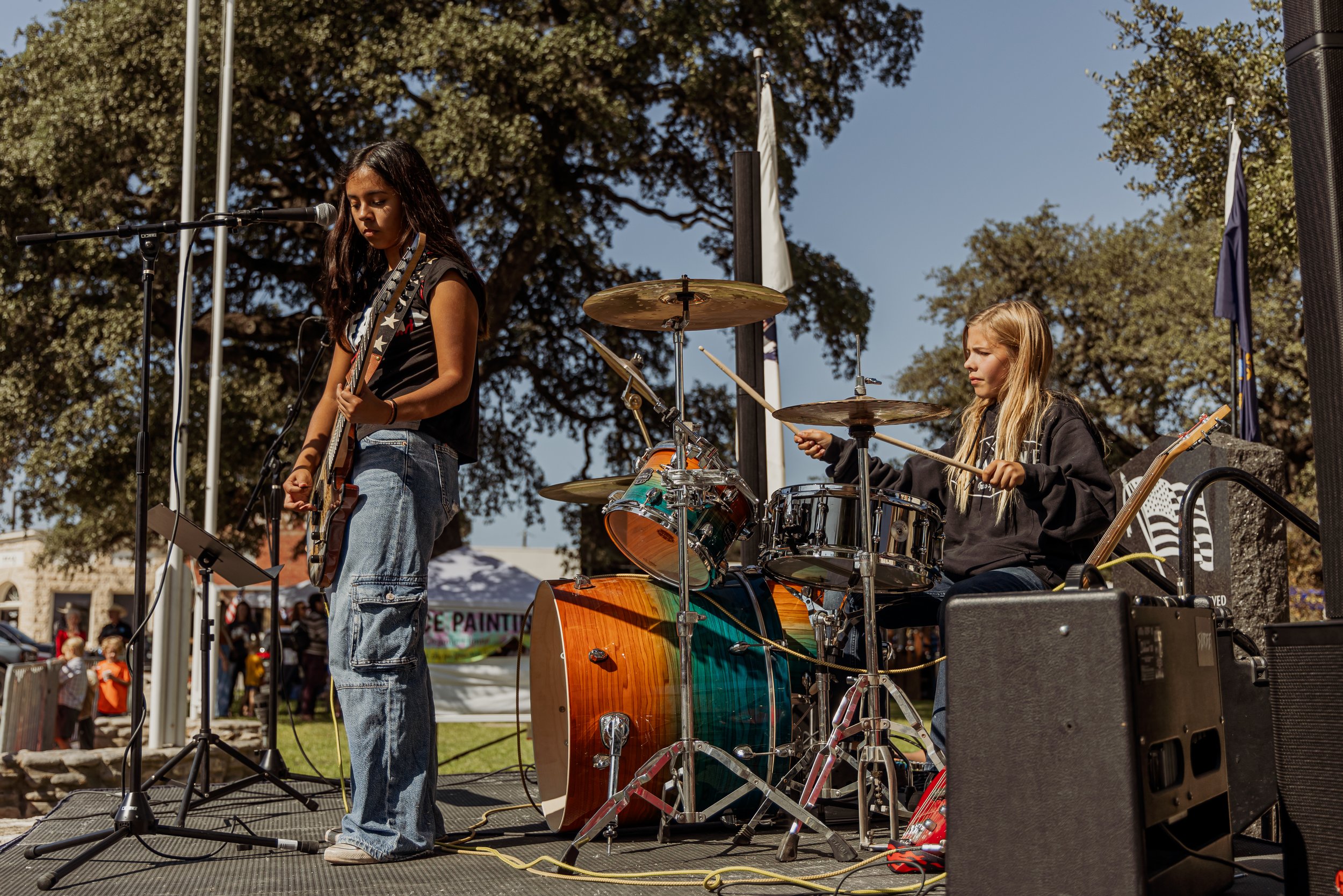 Two young girls playing musical instruments on an outdoor stage, one singing and playing guitar, the other on drums, with trees and a crowd in the background.