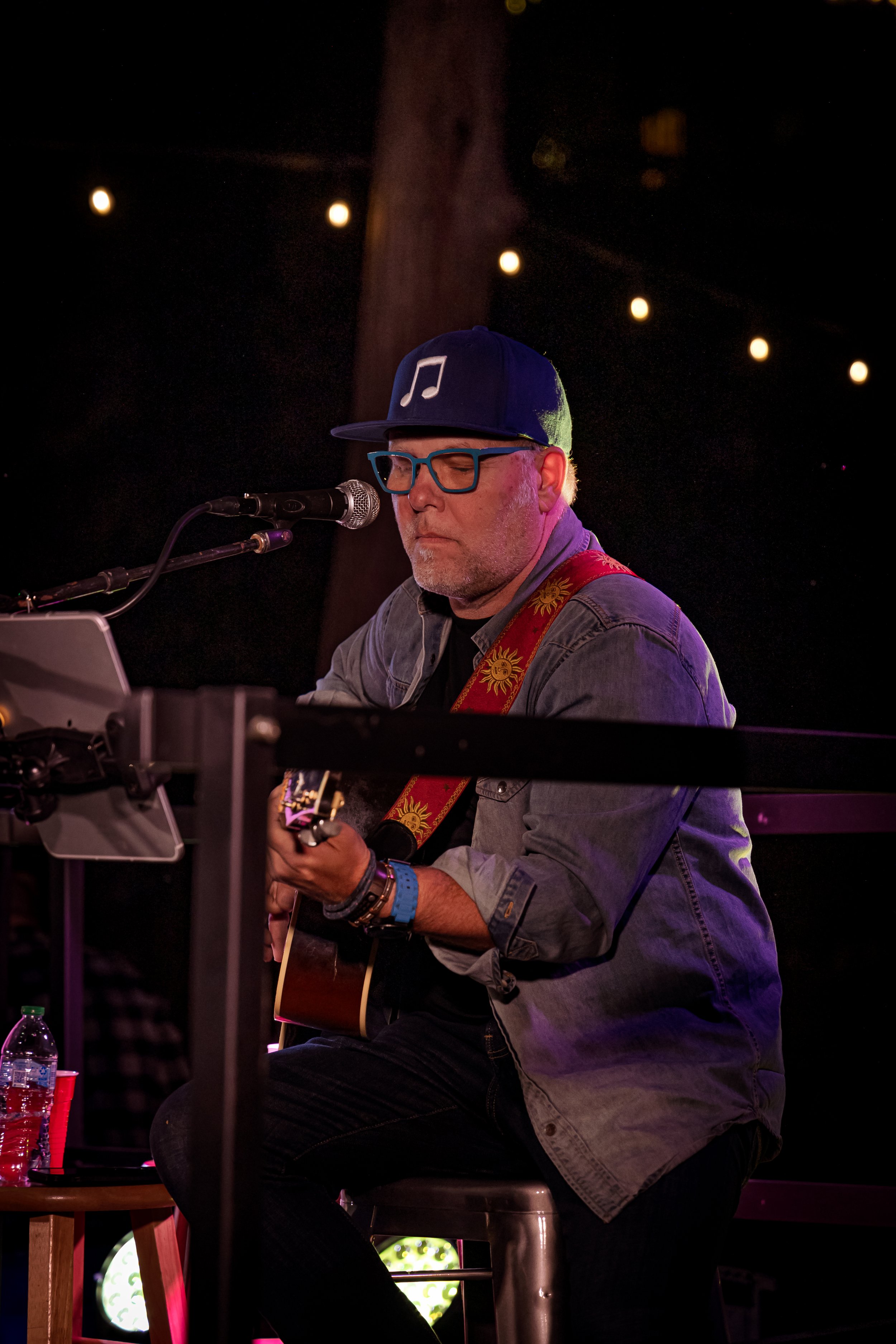 A man wearing a blue baseball cap with a musical note, glasses, and a gray denim jacket is sitting on a stool, playing an acoustic guitar and singing into a microphone. There is a water bottle and a red cup on a small table nearby, and string lights