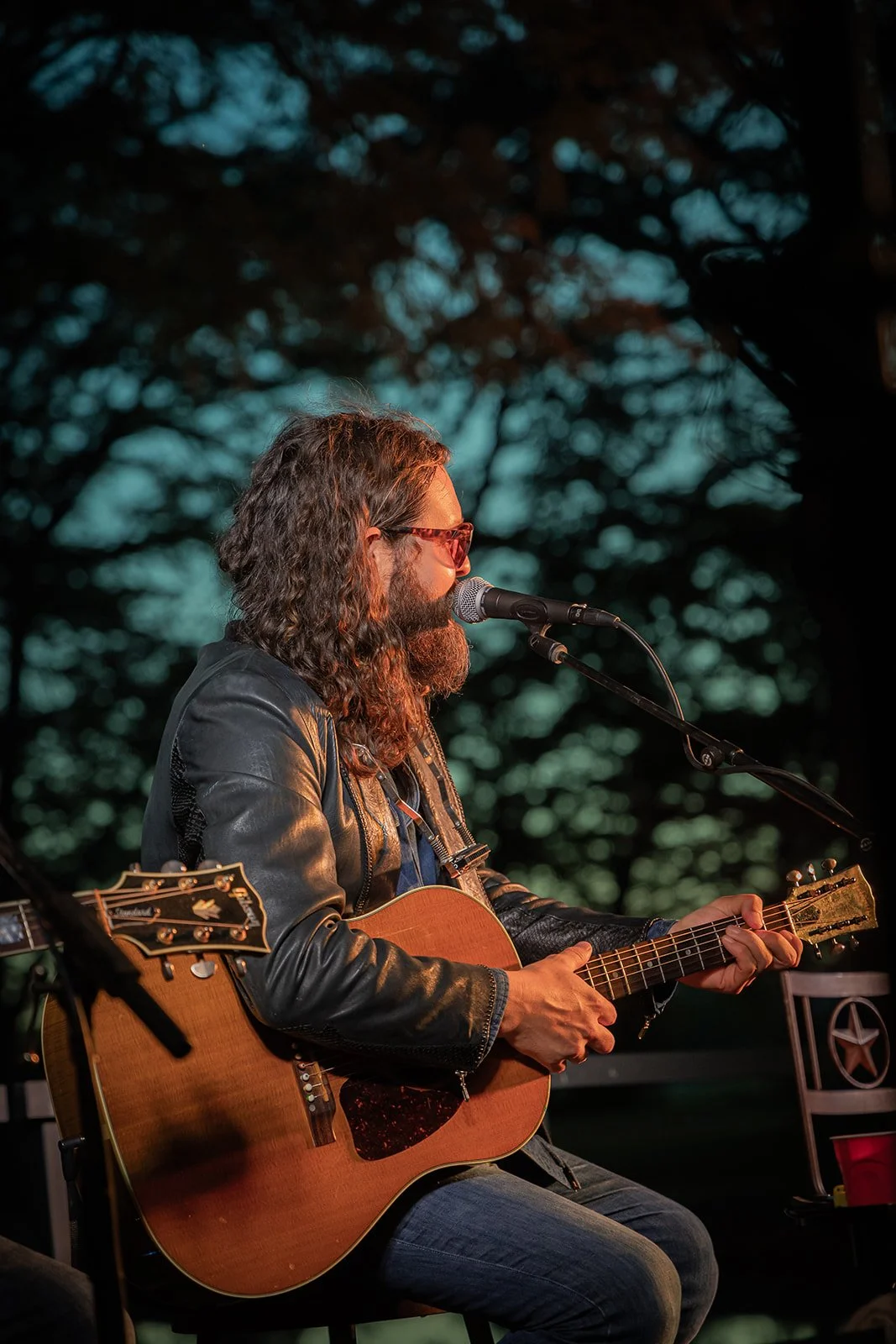 A man with long curly hair, a beard, and glasses, wearing a leather jacket, playing an acoustic guitar and singing into a microphone outdoors at dusk.