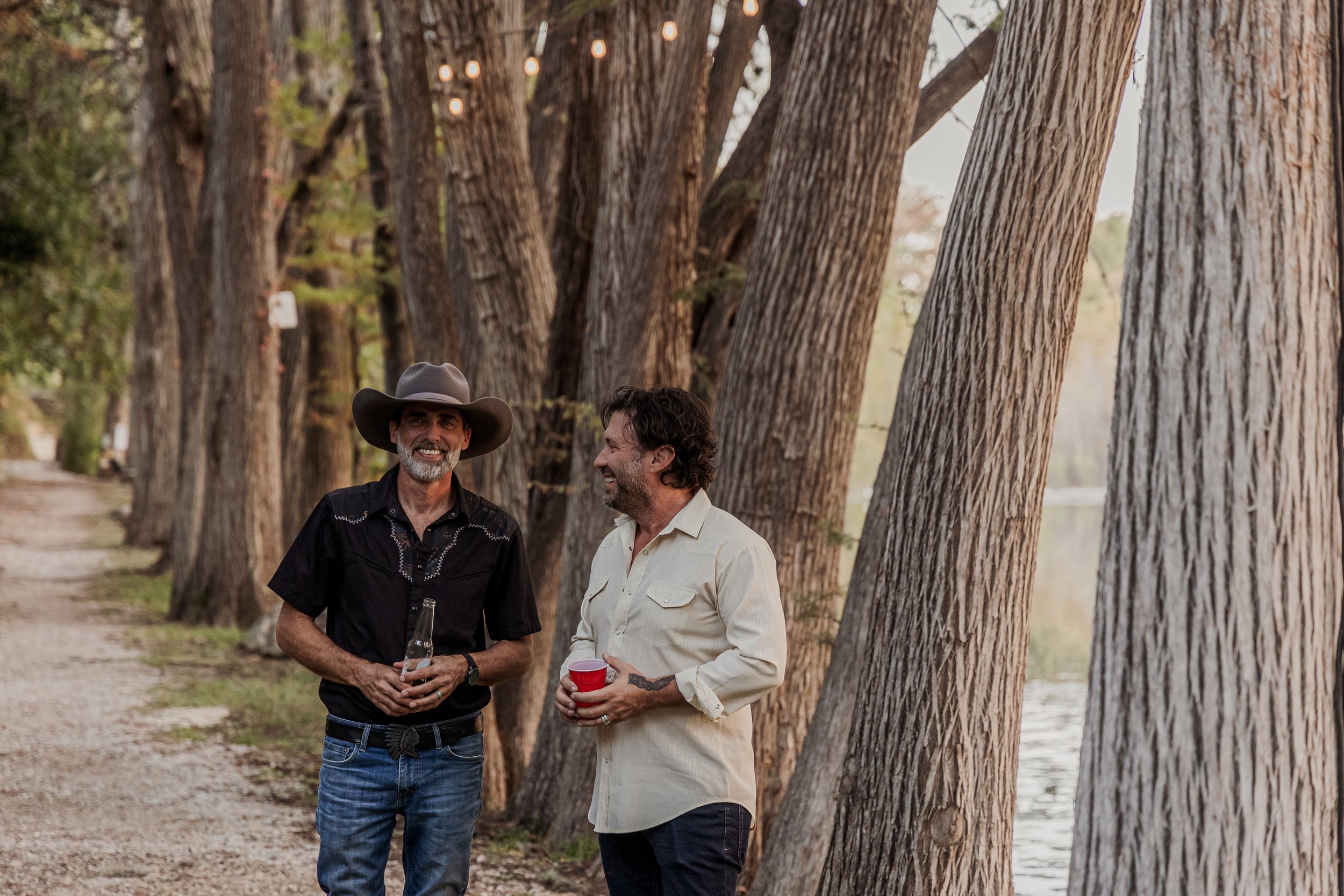 Two men are standing along a tree-lined path near a body of water, smiling and talking. One man wears a cowboy hat and a black shirt, holding a bottle. The other man wears a light-colored shirt and holds a red cup. It appears to be late afternoon or