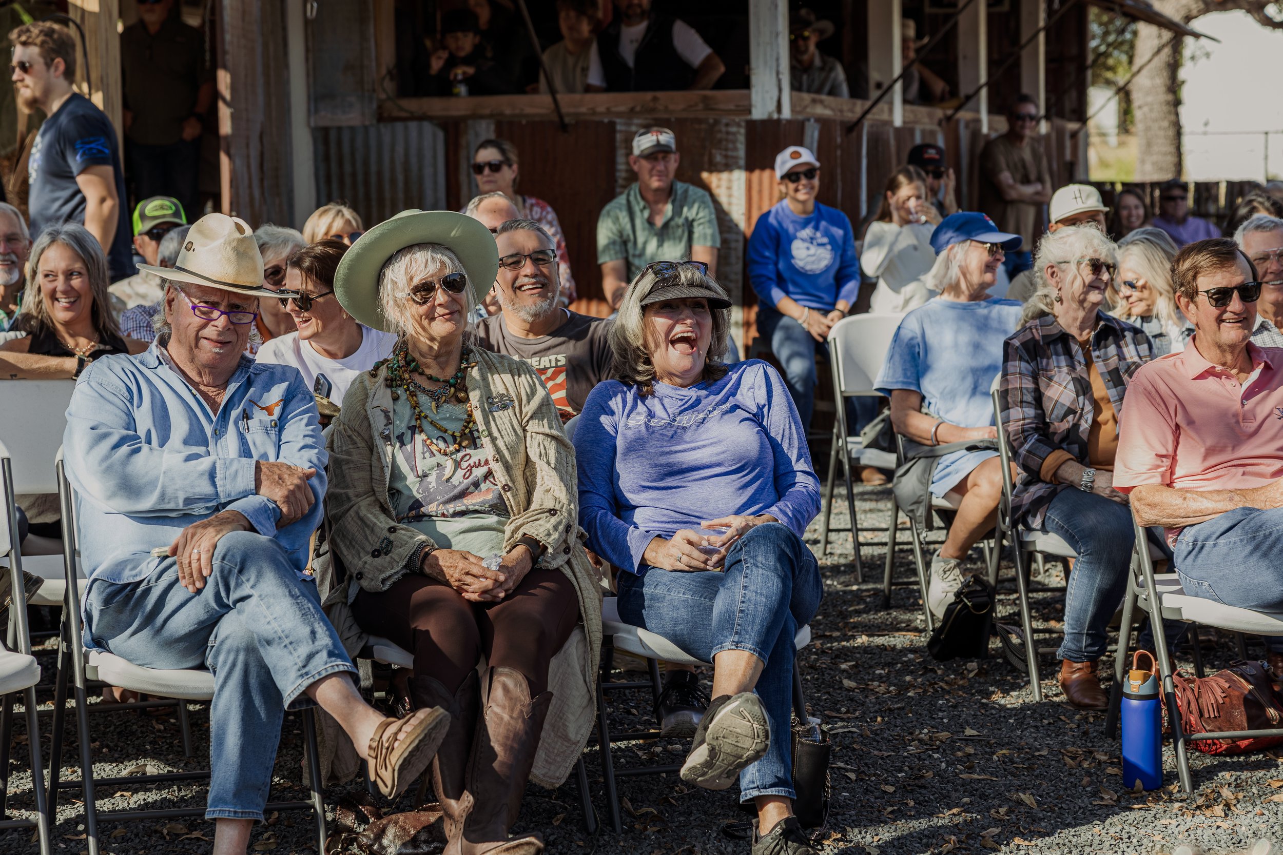A group of smiling people seated outdoors, attending an event with some wearing casual and country-style clothing, including hats, sunglasses, and jackets. The background shows a rustic wooden structure and more people sitting and standing, enjoying