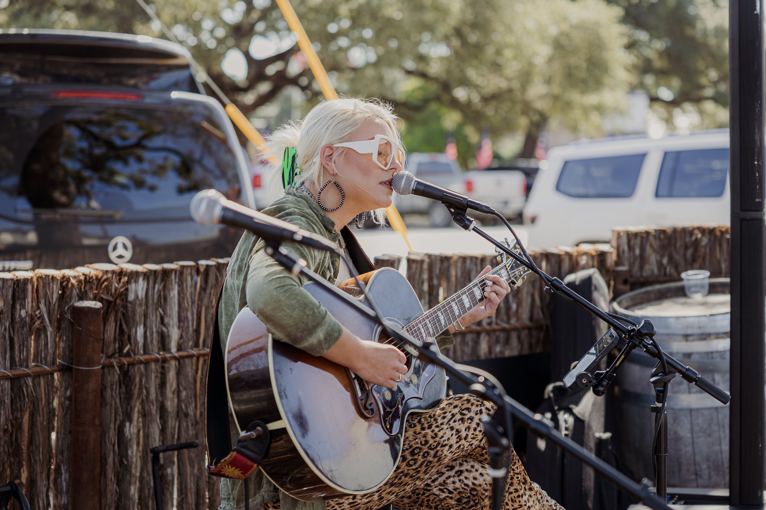 A woman with platinum blonde hair, wearing oversized white glasses, a green jacket, and leopard print pants, singing into a microphone while playing an acoustic guitar outdoors with parked cars and trees in the background.
