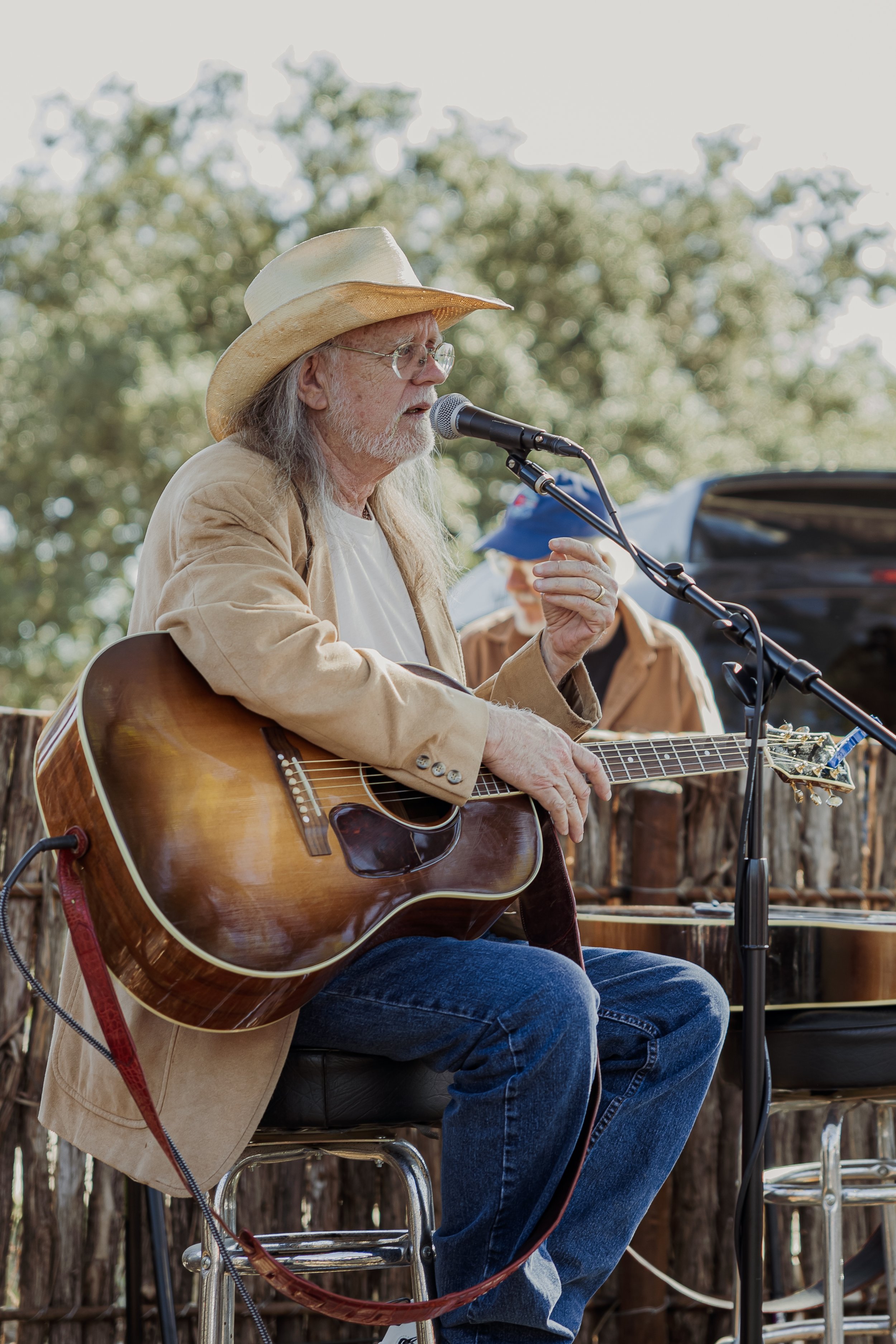 Older man with long gray hair, beard, glasses, wearing a tan blazer, white t-shirt, and cowboy hat, playing an acoustic guitar and singing into a microphone outdoors, with a person in a blue hat and brown jacket in the background.