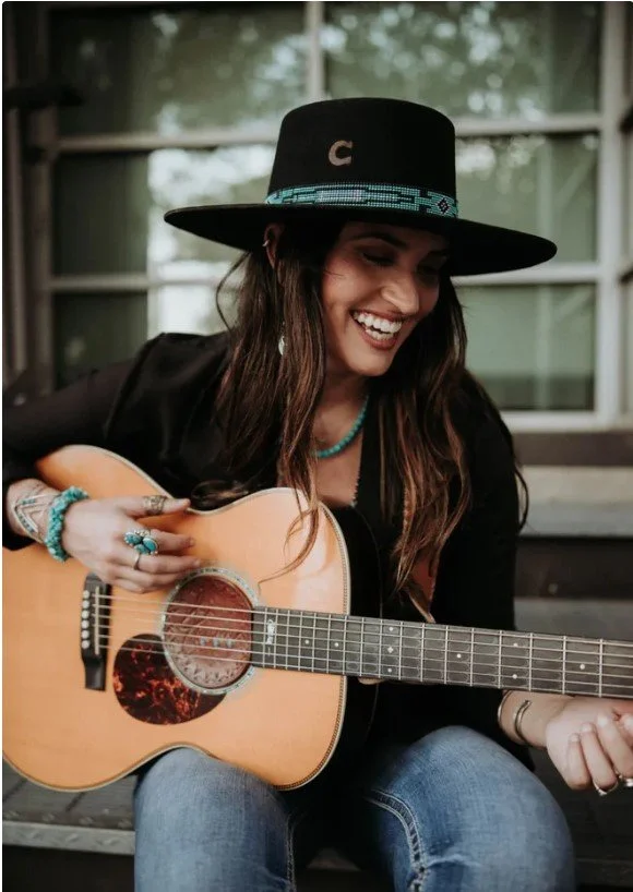 Young woman with long brown hair wearing a black hat and black jacket, smiling while playing an acoustic guitar, sitting outdoors near a house with large windows.