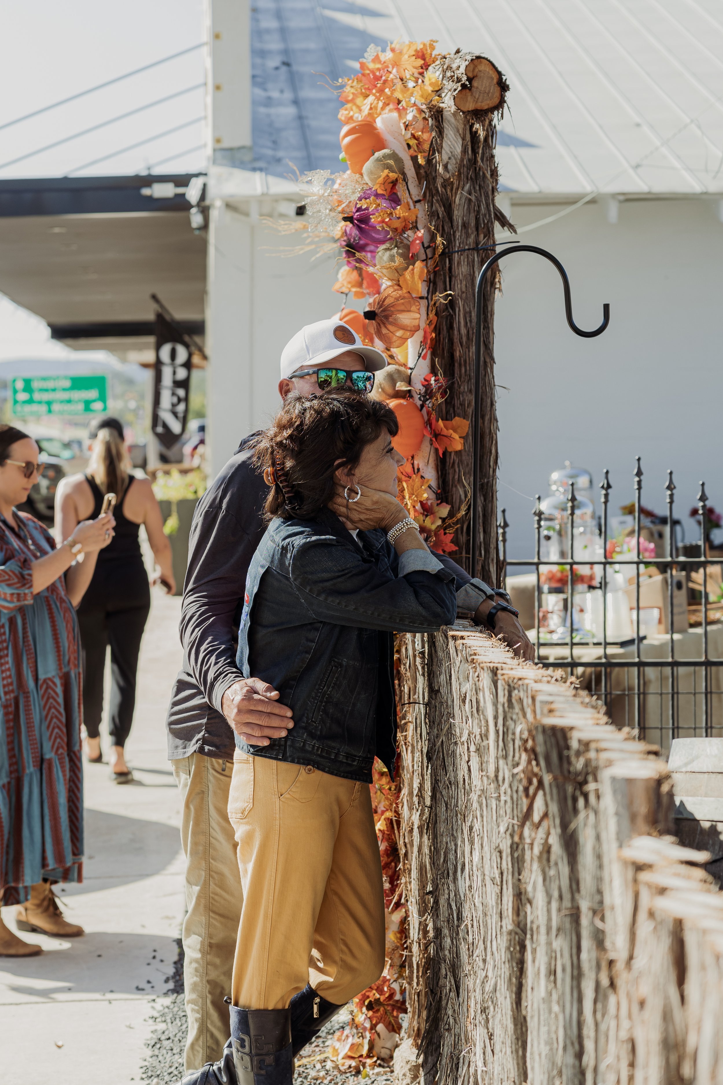 People standing outdoors near a decorated wooden post, some looking through a fence, with others taking photos.