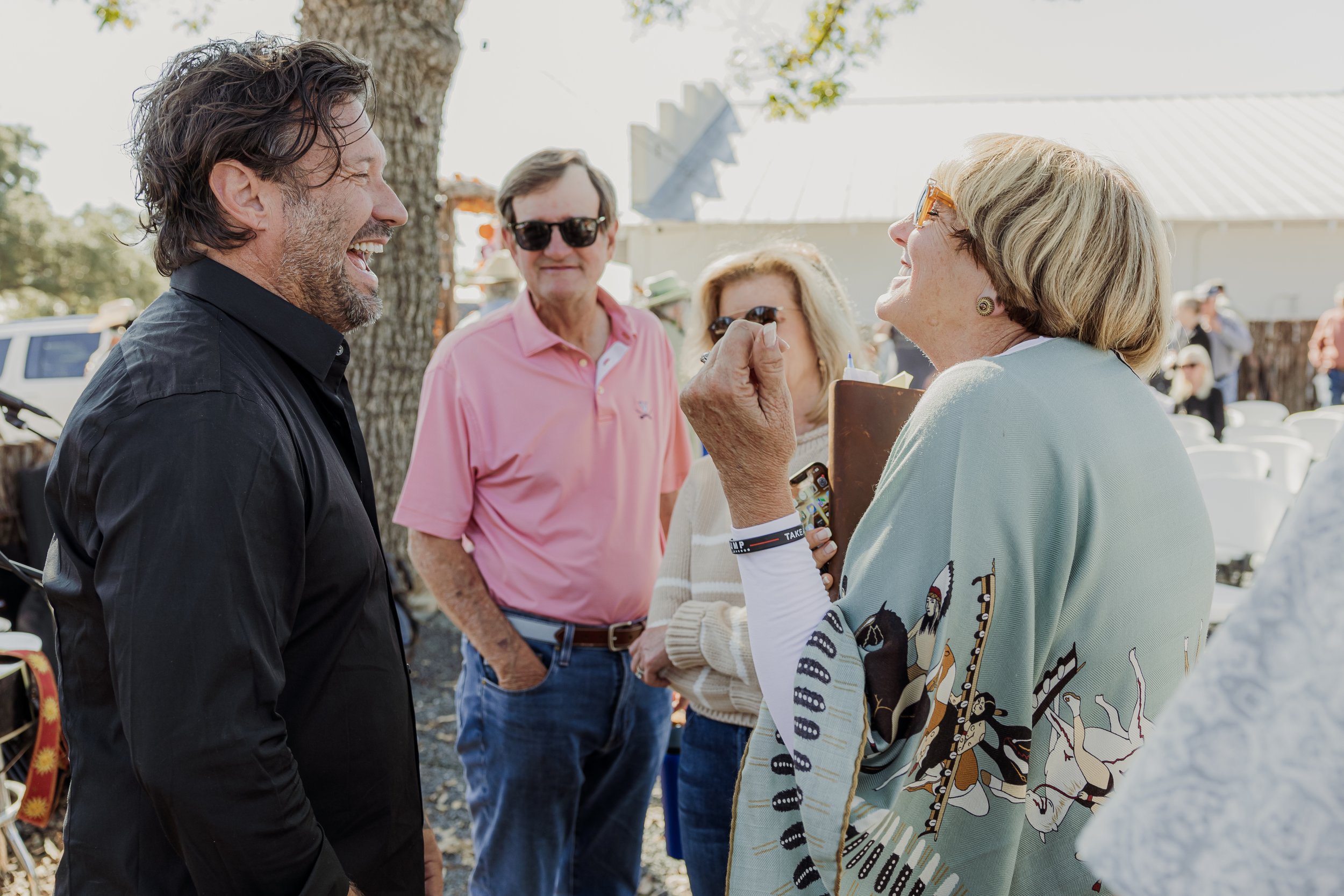 A man with dark, wavy hair and a beard laughing and talking to a woman with blond hair, glasses, and a colorful poncho, surrounded by three other people outdoors during daytime.