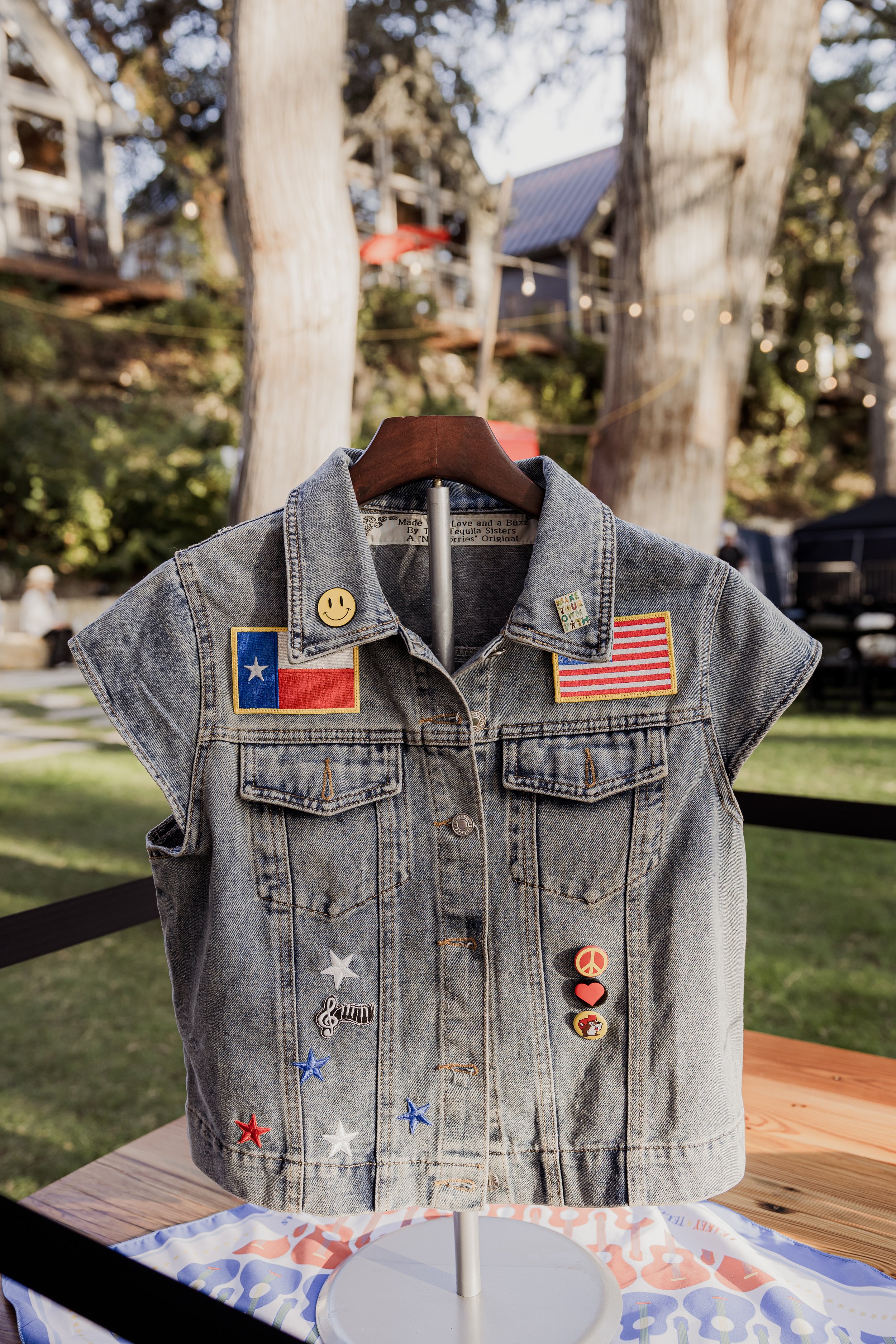 A sleeveless denim vest decorated with patriotic patches, smiley face button, and various colorful pins displayed on a stand outdoors with trees and buildings in the background.