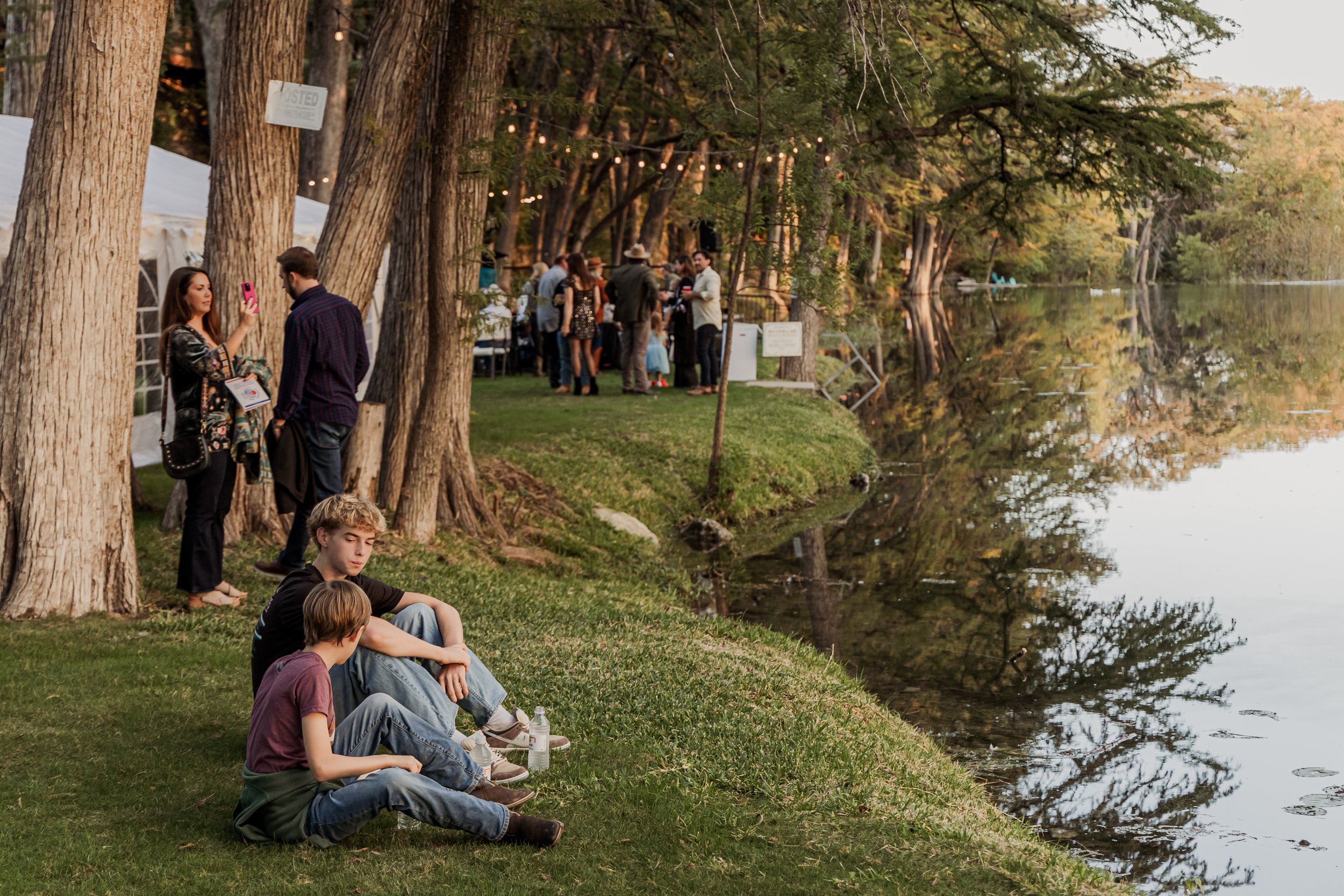 People gathered along a lakeside with trees, tents, and string lights, some sitting on the grass and others standing and chatting.
