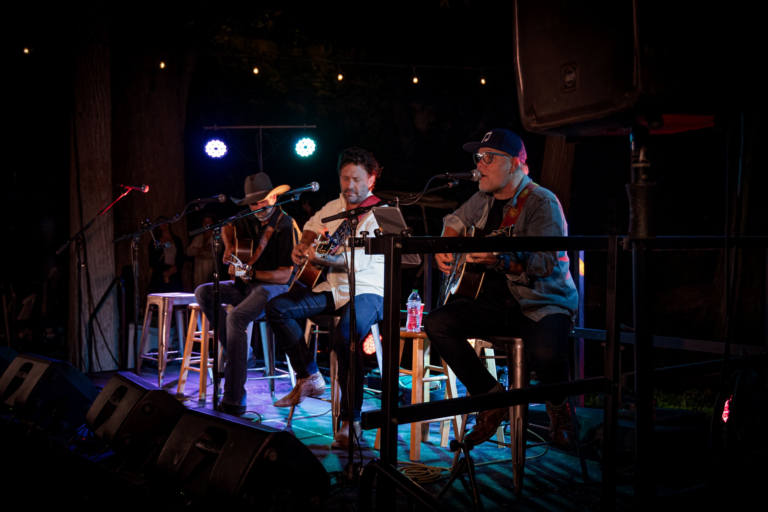 Three musicians performing on an outdoor stage at night, sitting on stools and playing acoustic guitars, with colored stage lights and tree trunks in the background.