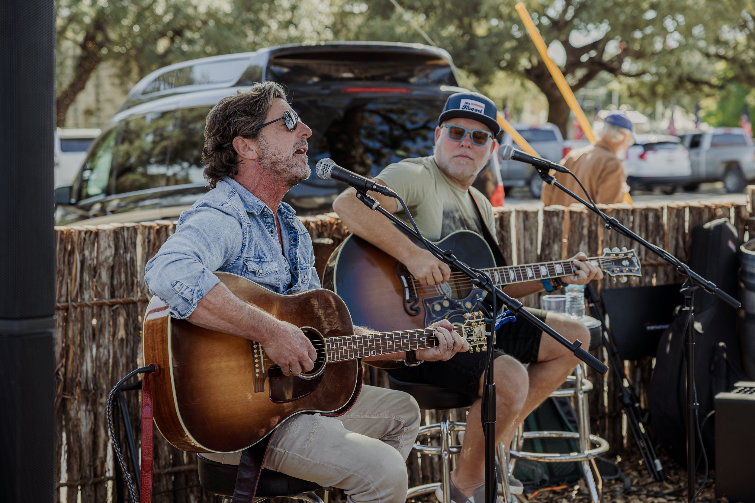 Two men playing acoustic guitars and singing outdoors at a live music event. The man on the left has gray hair, wears sunglasses, a denim shirt, and beige pants. The man on the right has a cap, sunglasses, a light green t-shirt, and black shorts. The